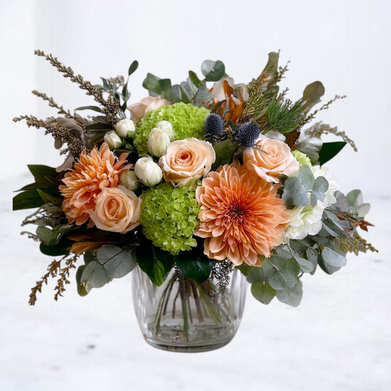 Gathered Grace Bouquet Gathered Grace Bouquet - Bring the beauty of the season indoors with our Gathered Grace Bouquet, a lush arrangement that blends warm peach dahlias, delicate roses, creamy ranunculus, green hydrangea, and striking blue thistle. Styled with seasonal foliage and textural accents, this bouquet captures the richness of fall in a modern garden-inspired design. Perfect for a thoughtful gift, a stunning centerpiece, or to add natural beauty to your home, this arrangement is both timeless and unique. Available for same-day flower delivery in Houston, Magnolia, The Woodlands, Spring, Conroe, Tomball, and surrounding areas, this bouquet makes an elegant choice for birthdays, anniversaries, or simply to brighten someone’s day.