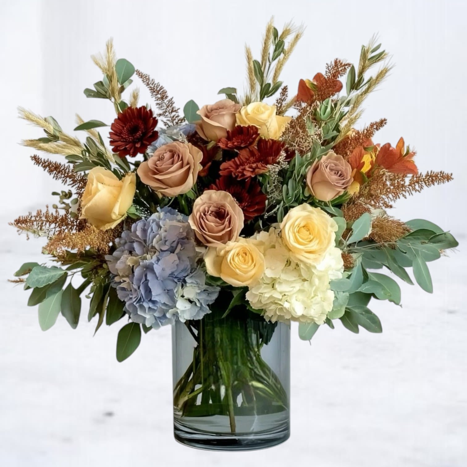 Harvest Meadow Bouquet Harvest Meadow Bouquet - The Harvest Meadow Bouquet captures the essence of autumn with a warm, rustic palette and natural textures. Golden yellow roses, rich rust mums, and soft taupe roses are paired with blue and ivory hydrangea, creating a unique balance of warmth and softness. Accented with golden aster, wheat stems, and fresh eucalyptus, this arrangement feels like a walk through a fall meadow. Designed in a clear glass vase, it makes a perfect seasonal centerpiece or thoughtful gift for Thanksgiving, birthdays, or simply celebrating autumn’s beauty. Available for same-day flower delivery in Houston and surrounding areas, this fall flower arrangement brings cozy charm to any occasion.