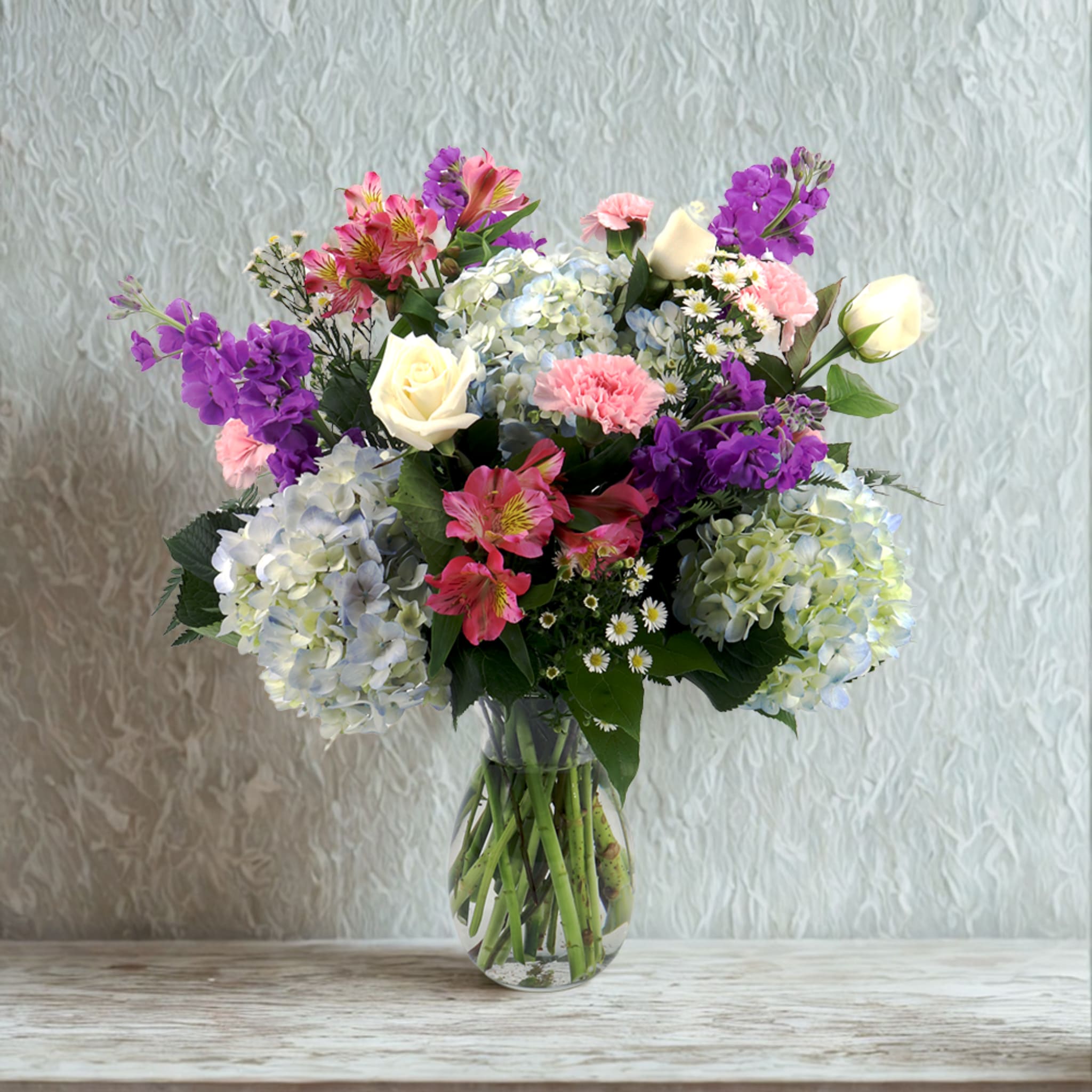 Gardens of Peace  - Gentle and restorative, Gardens of Peace was designed to bring a sense of calm and quiet beauty wherever it blooms. Blue hydrangea anchors the design with soft serenity, while stock and asters add airy texture and a whisper of movement. Roses and carnations lend classic warmth, complemented by the delicate charm of alstroemeria.  Together, these blooms create a tranquil palette that soothes the spirit and brightens the space — a floral reflection of balance, comfort, and grace.  Mindfully arranged at Birch &amp; Blossom, Gardens of Peace offers a moment of stillness and the soft reminder that beauty can bring peace.