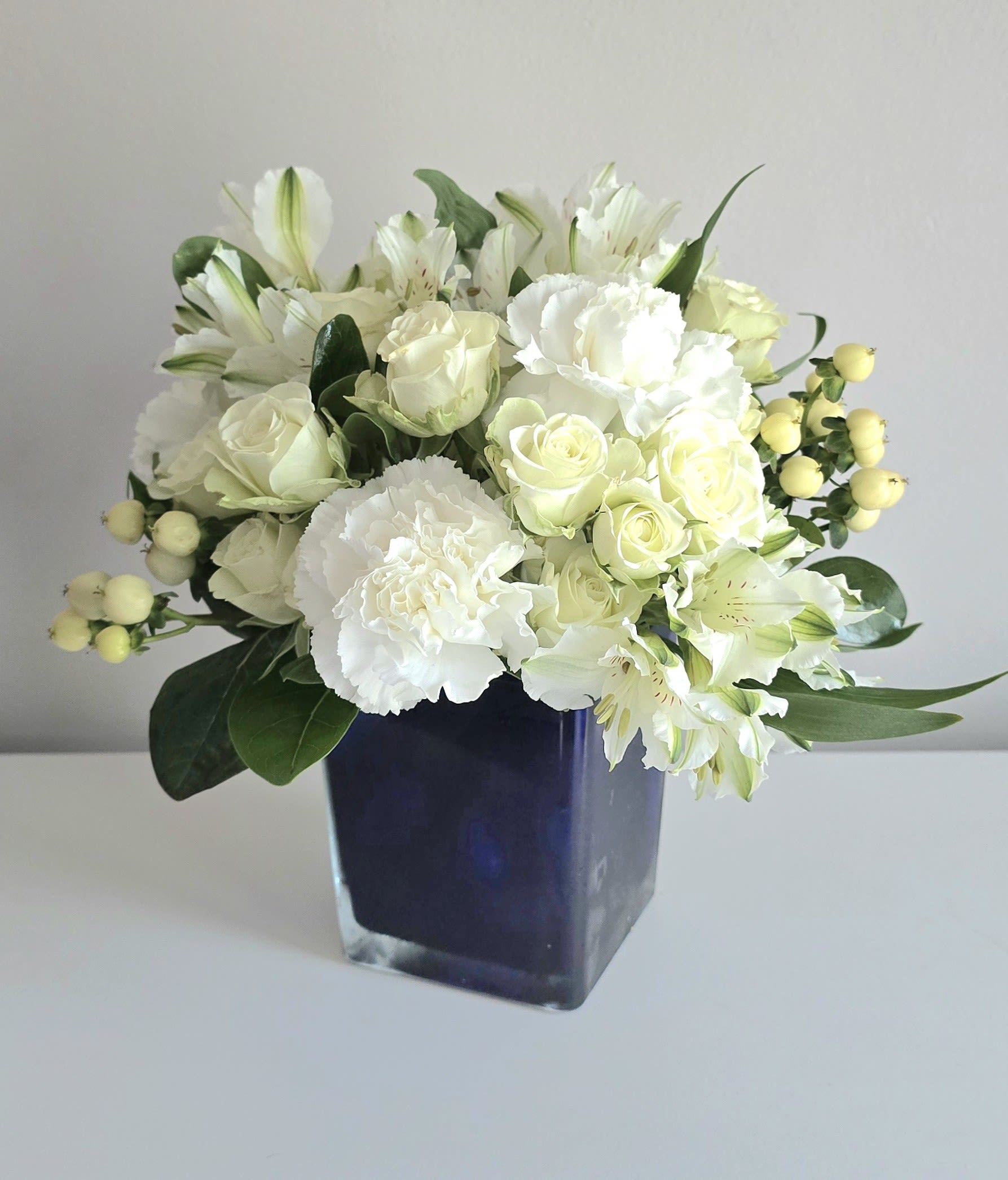 Sapphire Skies  - The freshest all white seasonal blooms, like white spray roses, berries, alstroemeria &amp; carnations fill a beautiful blue sapphire glass cube keepsake. Arranged low &amp; accented with dark greenery. Send this to friends &amp; families mourning the loss of a loved one.   Measures approx. 11&quot; tall &amp; 12&quot; wide Orientation: All around