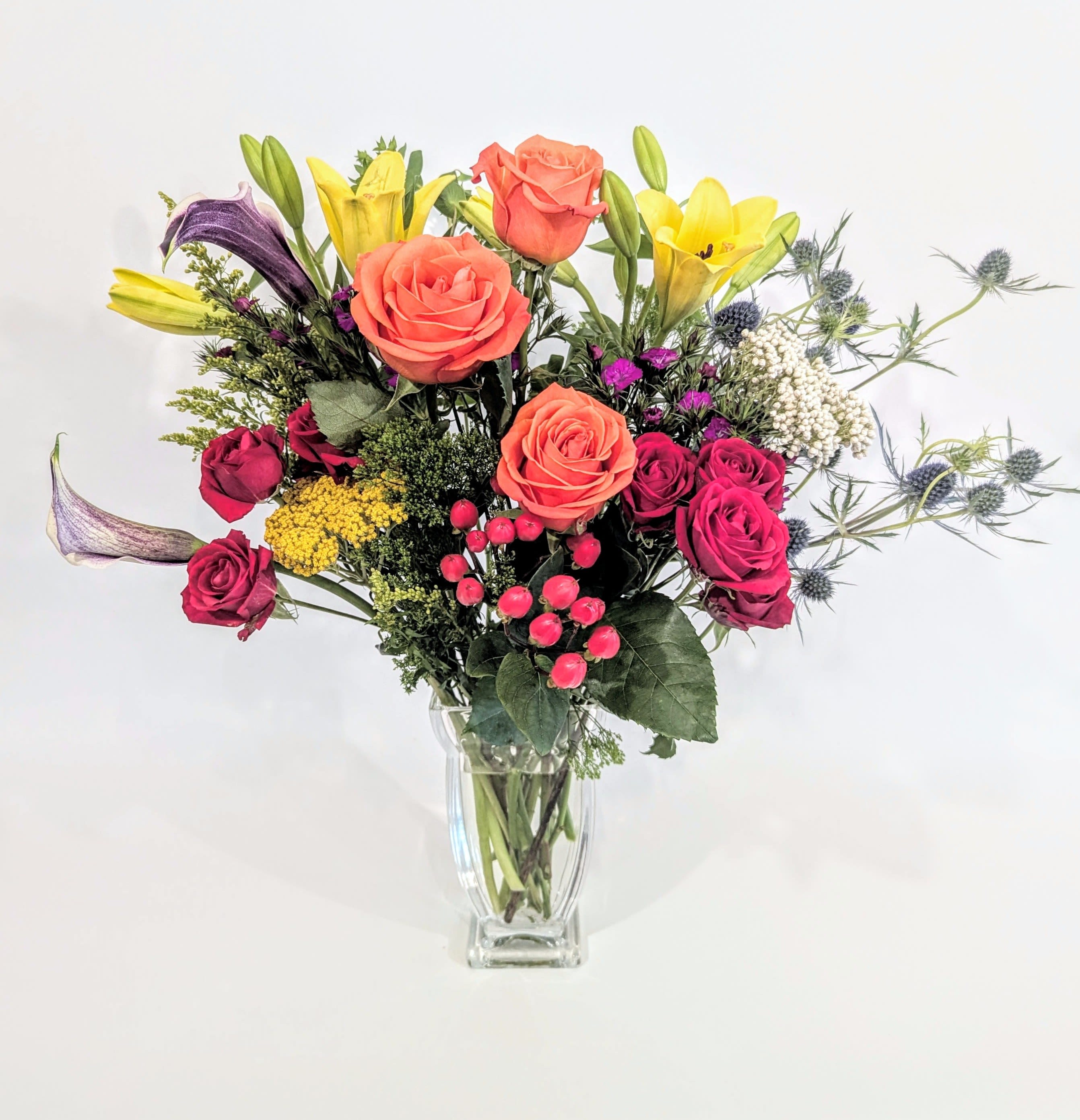 Fall In Love  - A beautiful and vibrant mixed bouquet featuring orange roses, purple calla lilies, red spray roses, yellow lilies, yarrow, rice flower, thistle, yellow solidago, purple sweet William, dianthus, red hypericum berries, bupleurum. Arranged natural and delivered in a high class glass couture vase. This arrangement is the perfect way to welcome in late summer and fall celebrations.   Measures approx. 20&quot; high and 17&quot; wide 