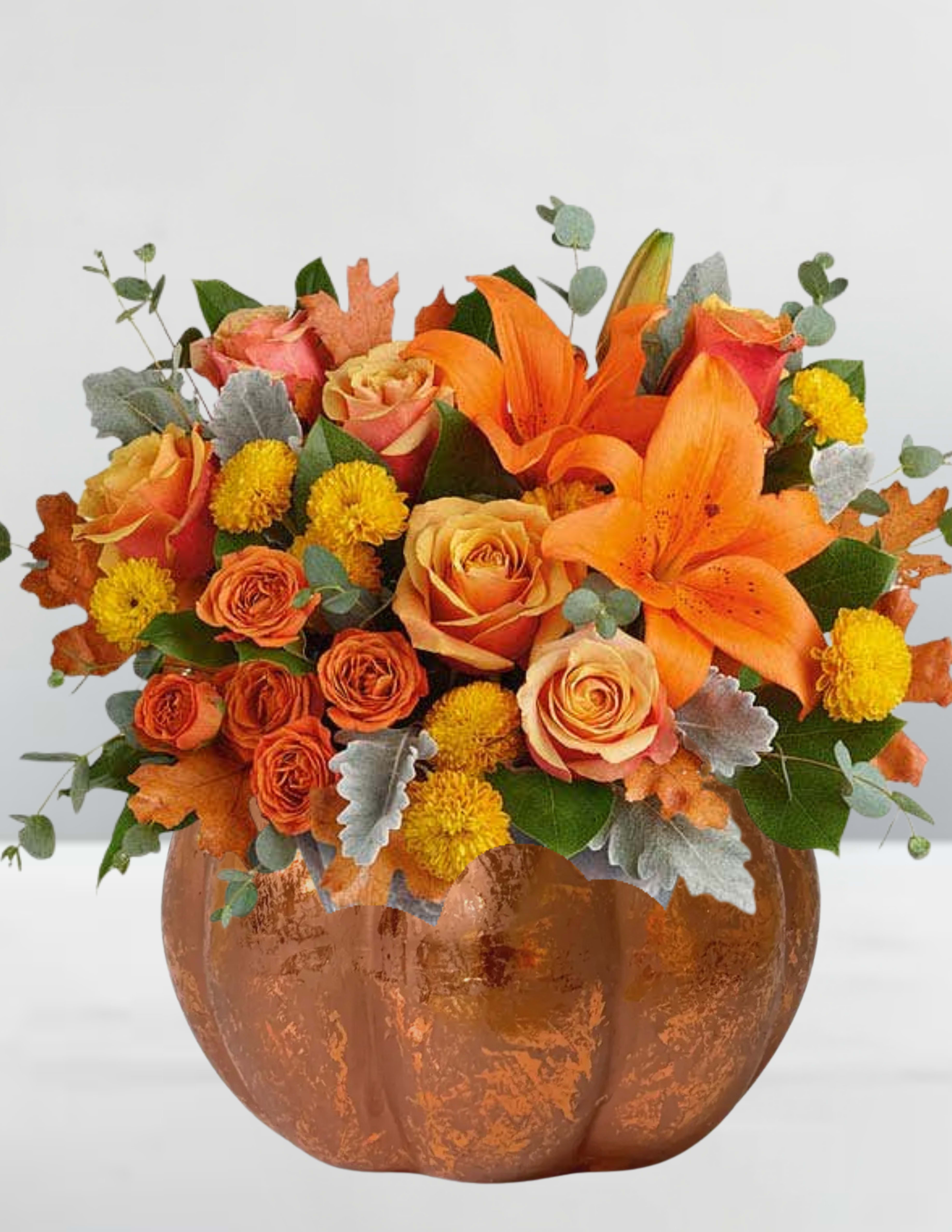 Harvest Glow Pumpkin Arrangement - Harvest Glow Pumpkin Arrangement  Celebrate the warmth and beauty of autumn with our Harvest Glow Pumpkin Arrangement. Nestled in a rustic ceramic pumpkin container, this radiant bouquet features vibrant orange lilies, golden yellow button mums, and a stunning variety of orange and peach roses. Accented with seasonal touches like faux autumn leaves, dusty miller, eucalyptus, and lush greenery, this arrangement brings the cozy charm of fall right into your home. Perfect for Thanksgiving centerpieces, fall birthdays, or seasonal décor, it’s a glowing reminder of harvest season’s natural splendor. 