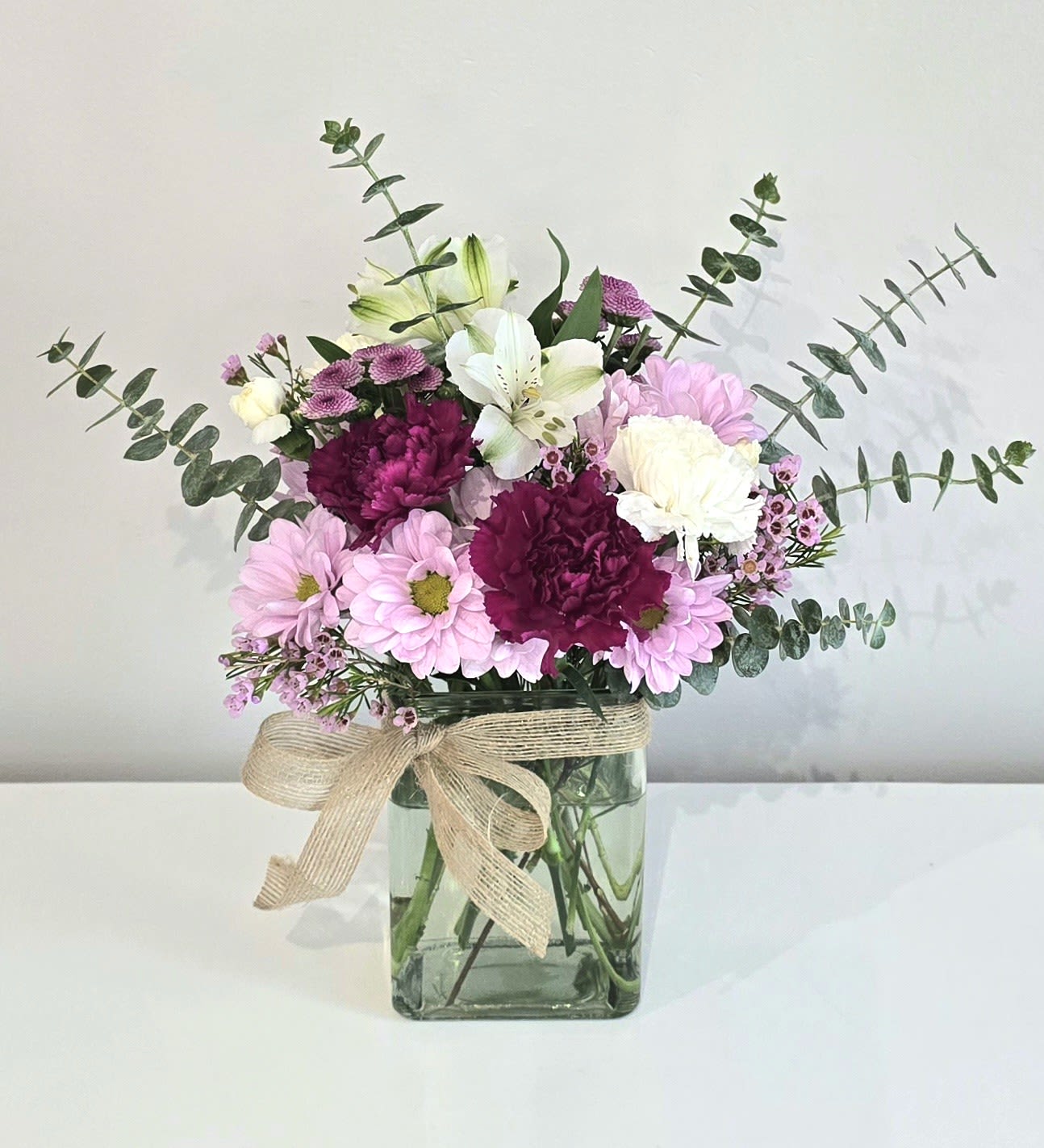 October Special - Petite Bouquet - This small but charming bouquet is just the right way to say I'm thinking of you. Lavender daisies, purple &amp; white carnations, alstroemeria, button spray poms, fragrant spiral eucalyptus &amp; waxflower are set inside a recycled glass rectangular vase &amp; accented with a pretty burlap bow. Send this for get well, sending thanks or just because occasions.   Available in STANDARD size only  Measures 13&quot; tall &amp; 13&quot; wide Orientation: Front focal