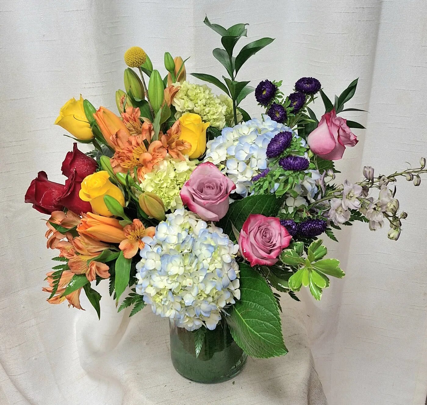 Passionate Petals (DL) - Introducing our Passionate Petals Bouquet, a bold and vibrant arrangement that bursts with a magnificent array of colors and blooms. This stunning bouquet features blue hydrangeas, red, purple, and yellow roses, blue delphinium, and orange lilies, all artfully arranged in a clear glass vase.  