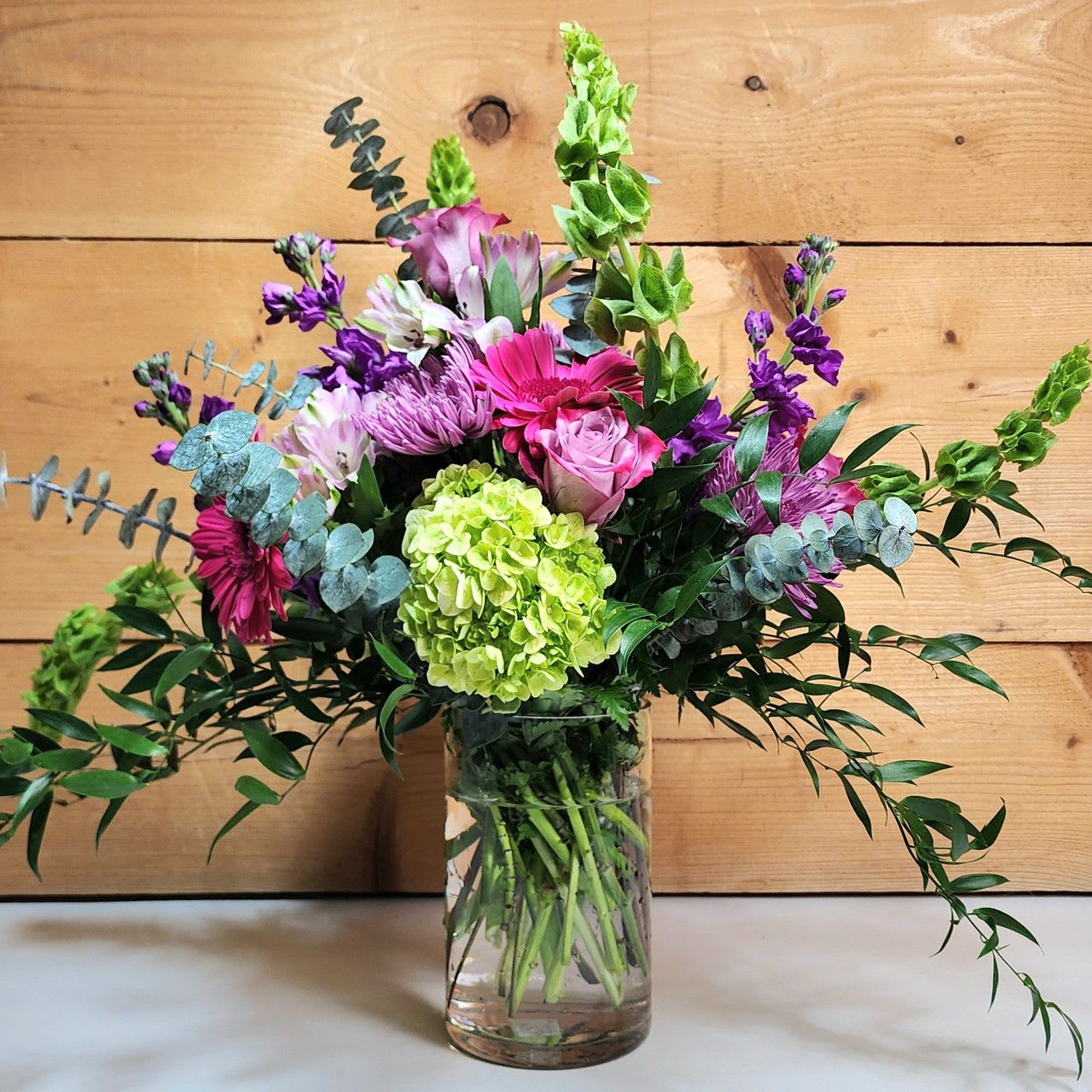 Majestic Harmony Majestic Harmony - Elevate any occasion with Majestic Harmony, a luxurious bouquet that blends the richness of purples and greens in an elegant display. This premium arrangement features lush purple stock, vibrant green hydrangea, and the velvety beauty of purple roses, creating a stunning foundation of color and texture. Dark pink gerbera add a playful pop, while the soft, ruffled petals of purple fujis bring a sense of sophistication. Lavender alstroemeria and delicate eucalyptus provide a soft, aromatic touch, with ruscus offering a refined, leafy contrast.    Displayed in a sleek glass vase, this arrangement exudes timeless beauty and grace, making it the perfect gift for someone truly special. Whether celebrating a milestone or simply expressing admiration, Majestic Harmony is an exquisite choice for those who appreciate the finer things in life.  approx 22" H x 22" W  PLEASE NOTE: Some flowers, vases and other products pictured are subject to availability, and may be substituted for a similar item if unavailable according to our substitution policy.