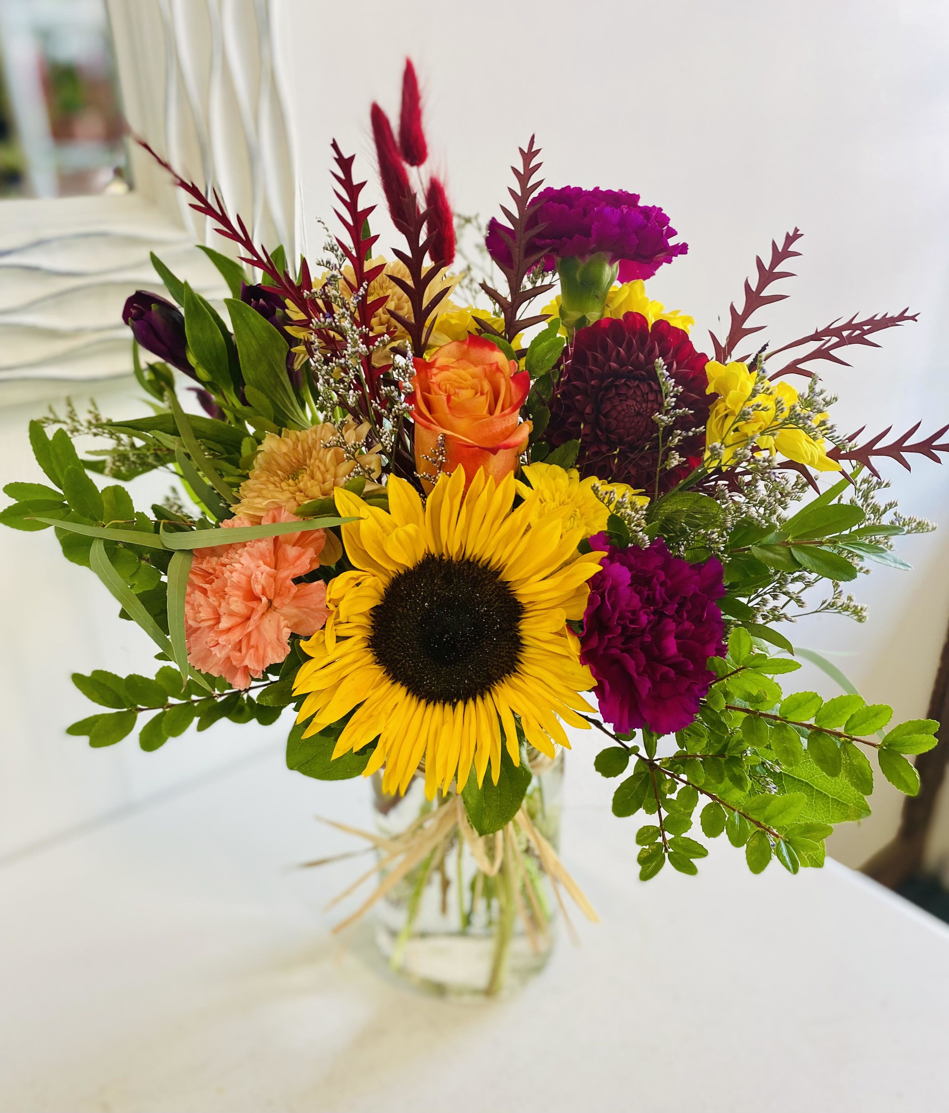 Sunflower Mason - Celebrate the season in full color with our Sunflower Mason arrangement — a cheerful blend of bold blooms and rustic charm. A radiant sunflower takes center stage, surrounded by vibrant orange roses, burgundy carnations, deep purple dahlias, sunny yellow daisies, and soft peach carnations. Accented with wisps of dried grasses, red foliage, and filler flowers, all gathered in a clear mason jar tied with raffia for a cozy, farmhouse finish.  This arrangement brings the feeling of a golden autumn day indoors — perfect for birthdays, thank-you gifts, or just because.