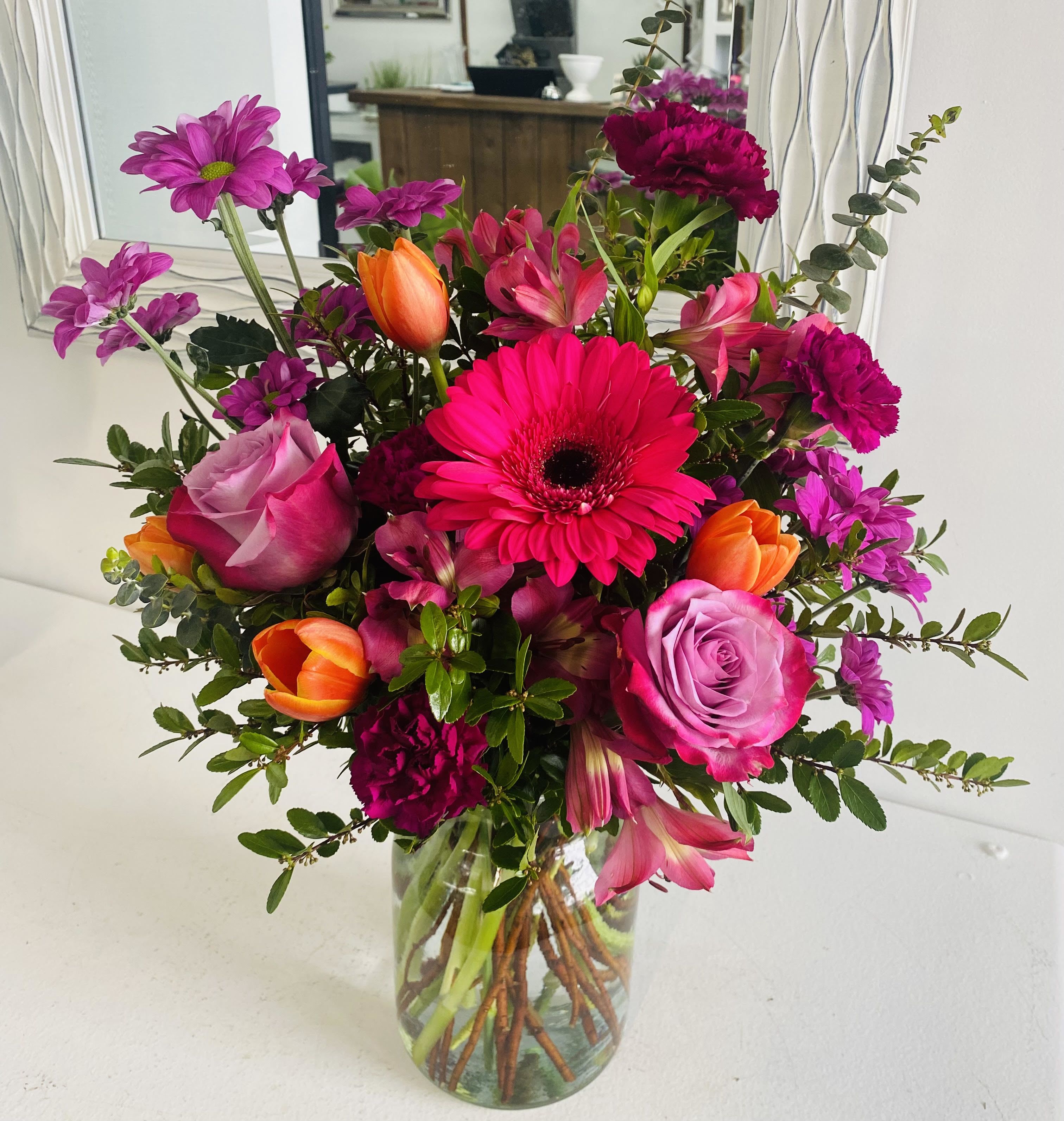 Sunscape Sunscape - Brighten any space with Sunscape — a stunning bouquet bursting with bold color and fresh energy. This eye-catching arrangement features vivid hot pink Gerbera daisies, elegant lavender roses, vibrant orange tulips, and rich magenta carnations, all artfully complemented by lush greenery and playful filler florals. Hand-designed in a clear glass vase, the Radiant Bloom is perfect for birthdays, celebrations, or simply bringing a burst of joy into any room. Its modern yet romantic palette makes it a versatile gift or centerpiece for any occasion.