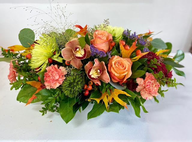 Fireside Florals Fireside Florals - This stunning fall centerpiece bursts with rich seasonal color and texture. Warm orange roses and peach carnations are paired with bronze cymbidium orchids, green spider mums, and deep burgundy accents. Bright berries, autumn leaves, and lush greenery complete the look, bringing the beauty of the harvest season indoors. Designed low and full, it’s the perfect arrangement for a dining table, Thanksgiving celebration, or to add a cozy autumn touch to any space.