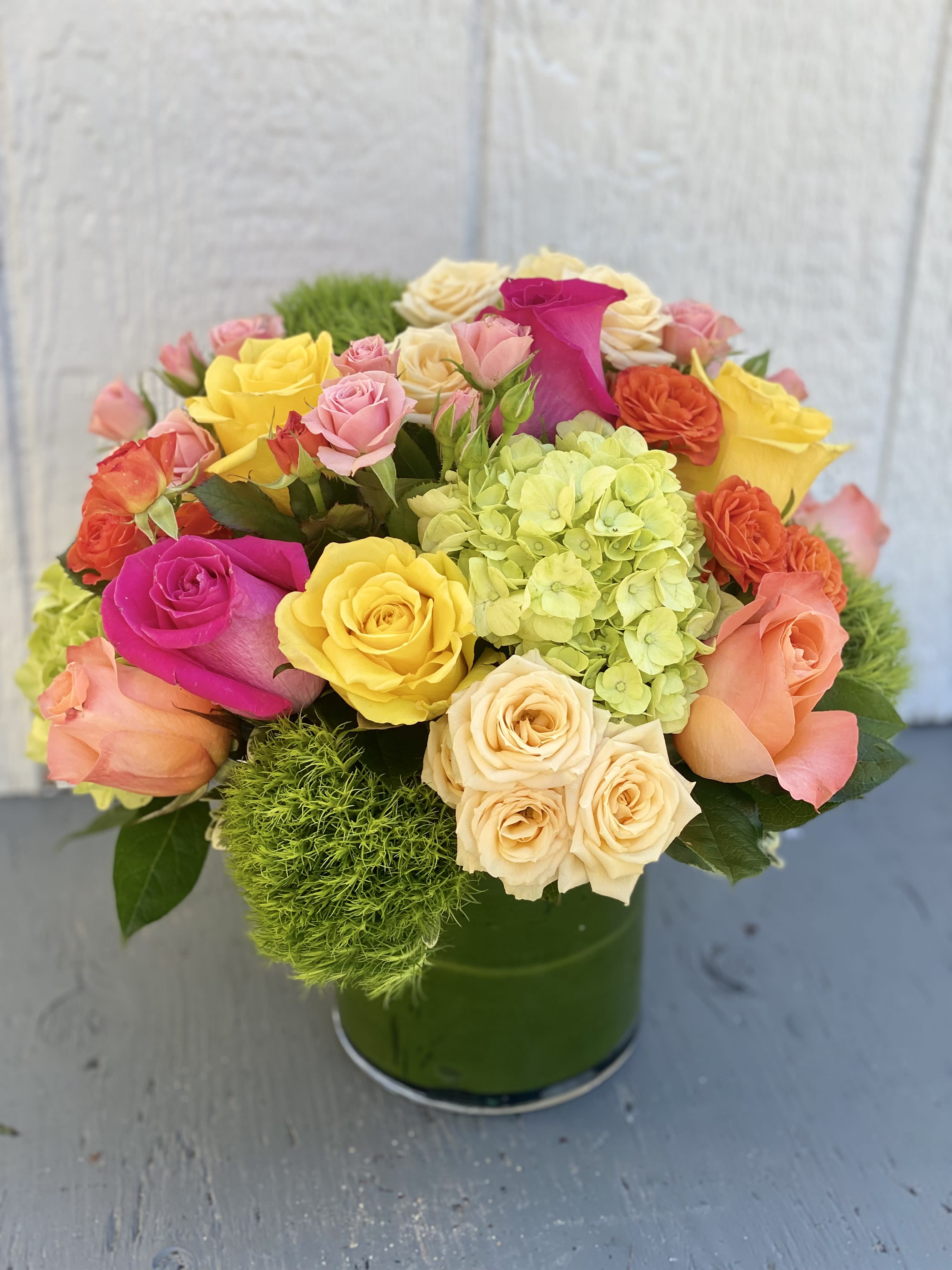 Blossoms in Vogue  - Blow someone away with a bouquet that has so much style all it needs is its own runway.  Brilliant green hydrangea, hot pink, orange, and yellow roses with complementary spray roses and greens are hand-delivered in stunning style: a wide cylinder vase that's been elegantly lined with leaves. This bouquet is a singular sensation! Deluxe size shown in photo  Approximately 13&quot; W x 12&quot; H  Orientation: All-Around      As Shown : T74-1A     Deluxe : T74-1B     Premium : T74-1C  