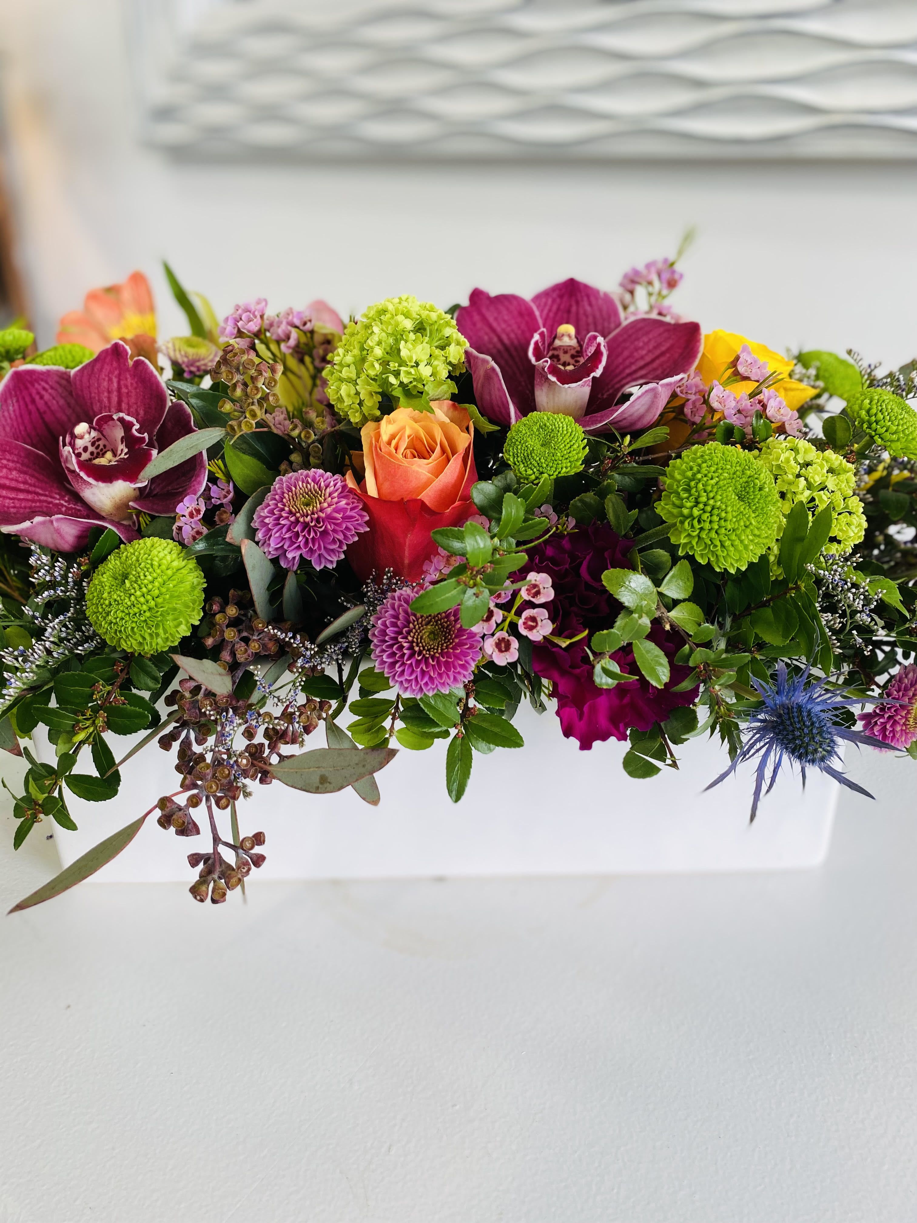 Window Box Blooms Window Box Blooms - This bold and colorful arrangement bursts with energy and texture, featuring rich burgundy orchids, vibrant orange and yellow roses, chartreuse hydrangea, playful button mums, and lavender dahlias. Accented with waxflower, eucalyptus, and touches of thistle, all artfully designed in a sleek white ceramic container, it’s a modern and joyful statement piece for any celebration.