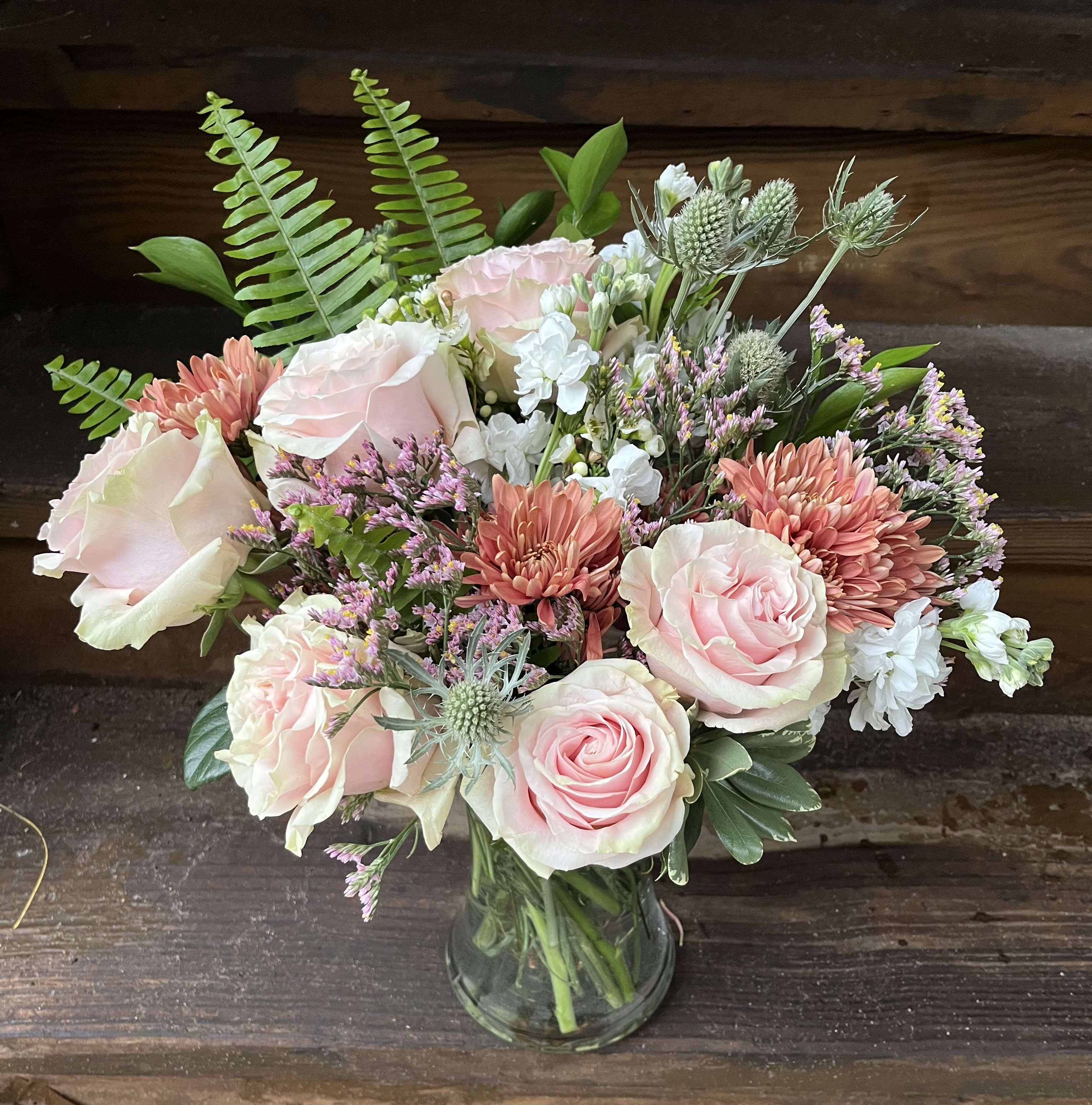 Amour Amour - Lovely Open Light Pink Garden Roses with Terra Cotta Mums, White Stock, Light Pink Limonlium, and a variety of fun Green Accents. Ideal for a birthday, anniversary or just because. Front Facing Bouquet in a 8" glass vase.