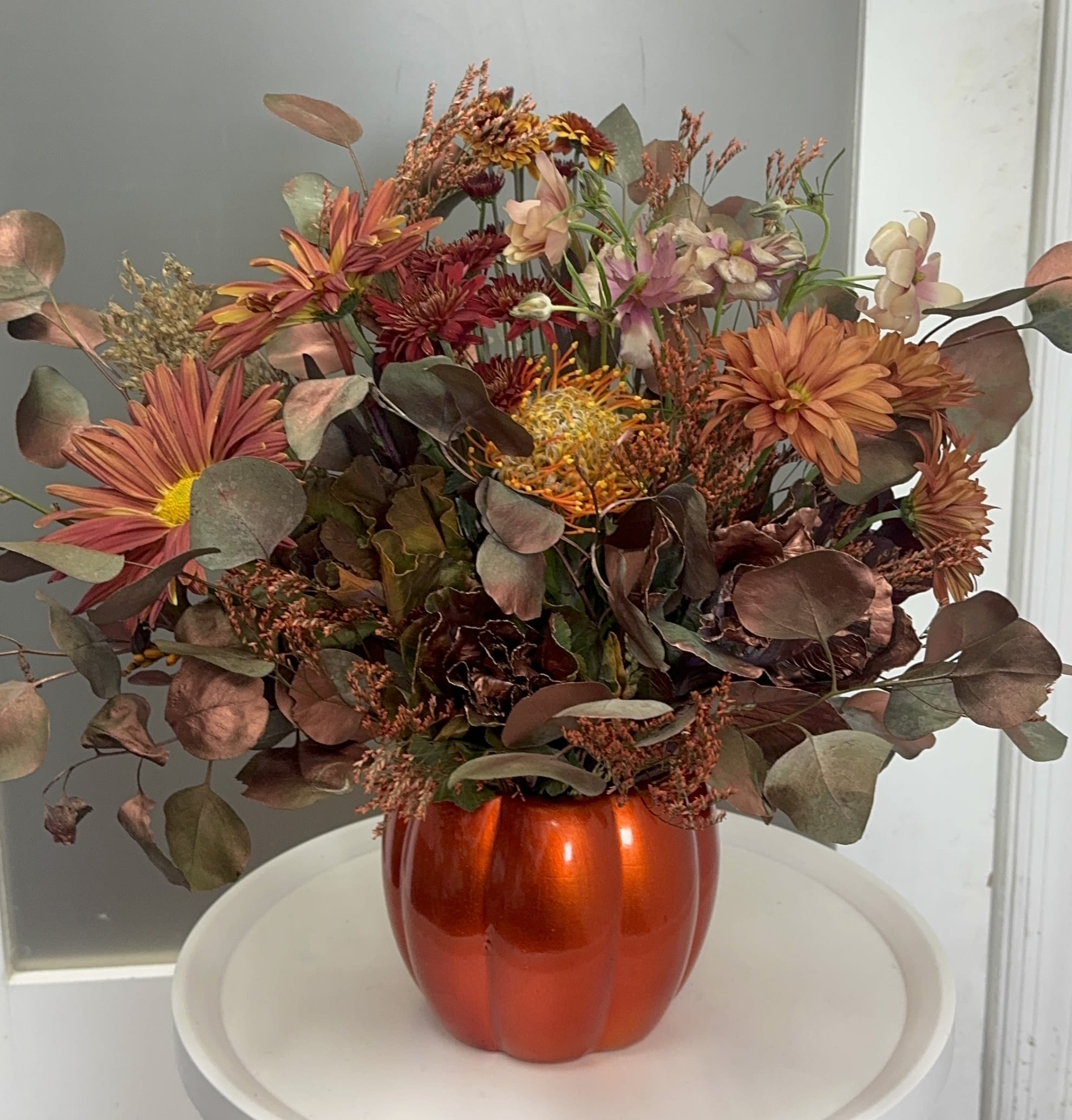 Autumn Glow  - Una fusión de texturas y colores que evocan la magia del otoño. Brillo de Otoño combina flores en tonos cálidos —terracota, cobre y dorado— en una elegante base con forma de calabaza metálica. Ideal para decorar espacios acogedores o regalar gratitud en temporada de Thanksgiving.