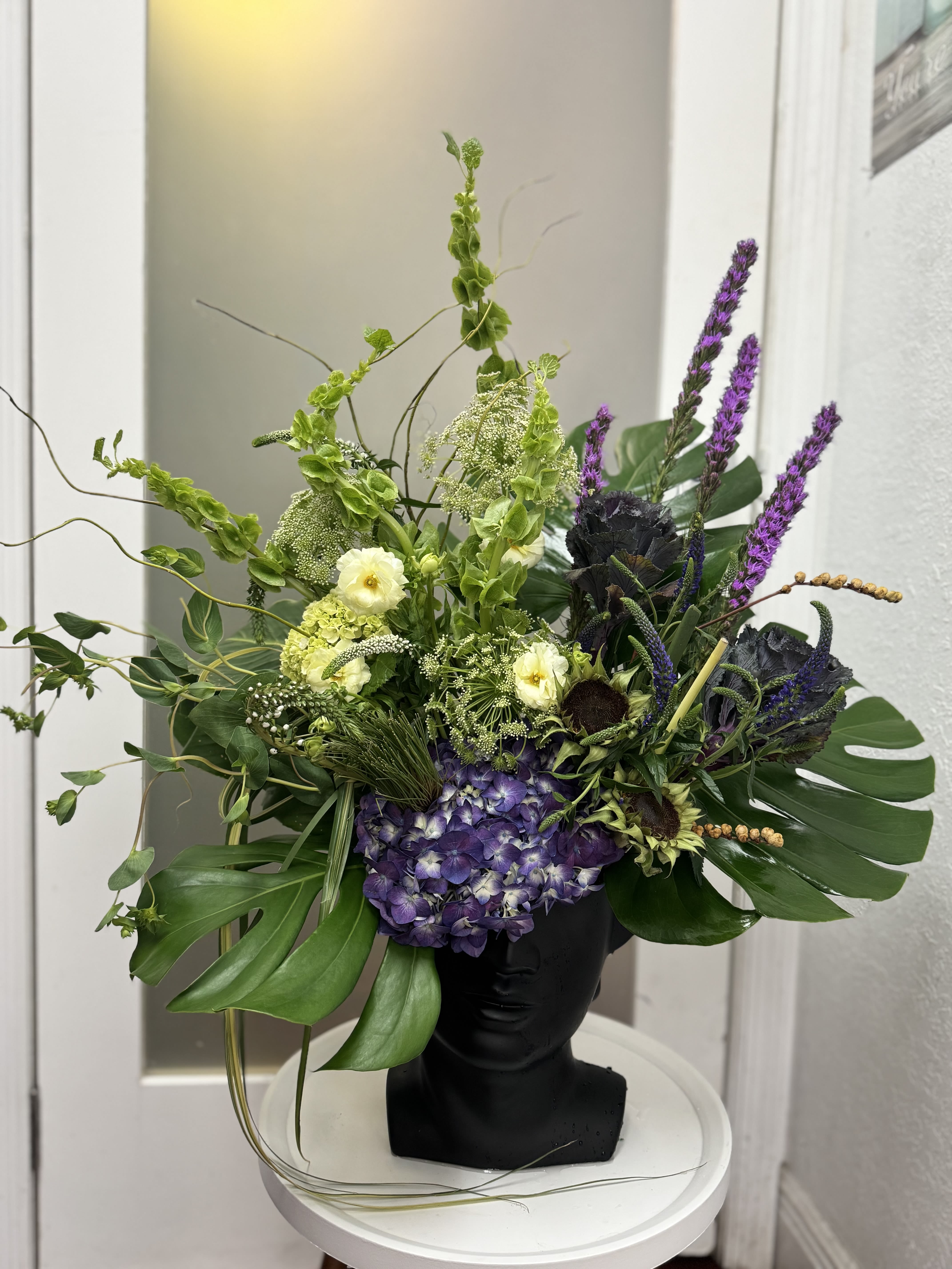 Green Muse - A design inspired by the harmony between nature and art. Green Muse blends shades of green and violet in a sculptural composition set on a head-shaped vase. Featuring hydrangeas, bells of Ireland, liatris, and tropical foliage, it embodies a perfect balance of elegance and artistic expression. Perfect for modern interiors, creative gifts, or events that celebrate art and individuality.