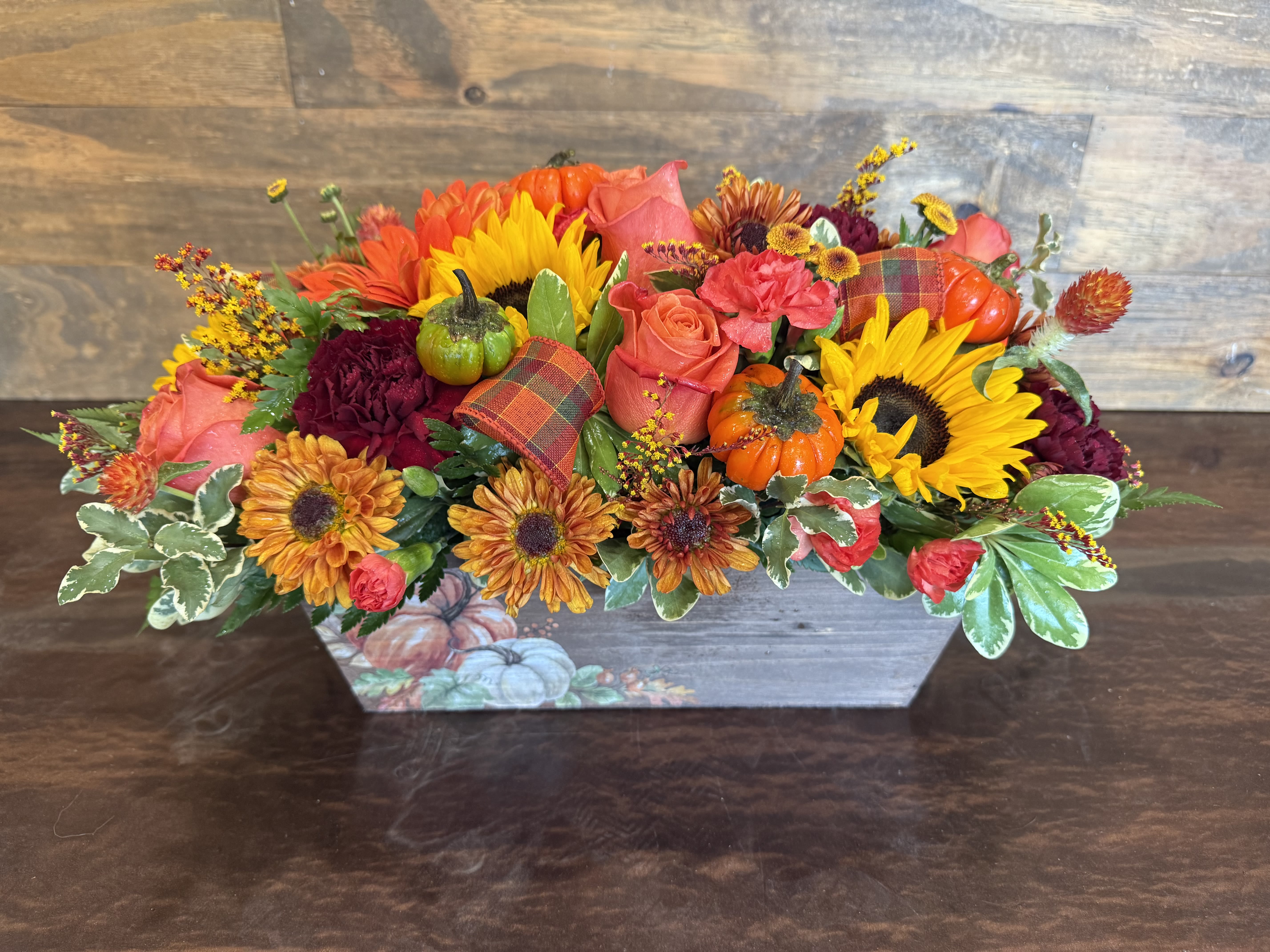 Autumn Gathering  - Celebrate the season of gratitude with this vibrant  arrangement overflowing with autumn charm. Bursting with golden sunflowers, rich burgundy blooms, and warm orange roses, it's accented by miniature pumpkins, plaid ribbon, and lush seasonal greens. Designed in a rustic wooden container, this arrangement brings the cozy glow of harvest gatherings to your holiday table. Perfect for Thanksgiving feasts, hostess gifts, or simply filling your home with the spirit of the season.