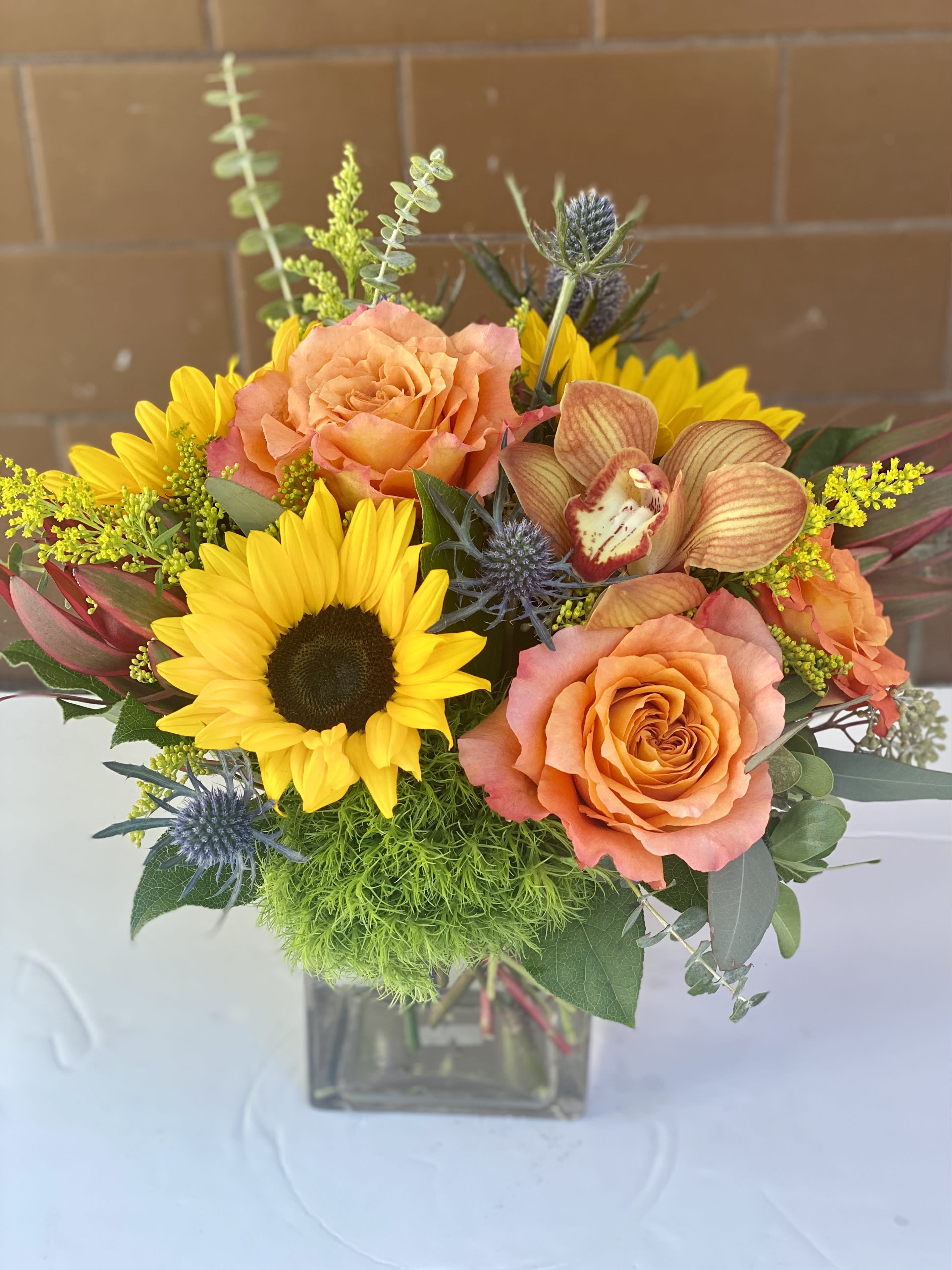 Petite Autumn Cube - A delightful cube bouquet. We start with a quality glass cube vase filled with long lasting and festive fall colors arranged and delivered for you. This bouquet has sunflowers, roses, cymbidium orchid and roses. This is a very trendy arrangement that may be used as a centerpiece, home decor piece or as a welcome hostess gift.
