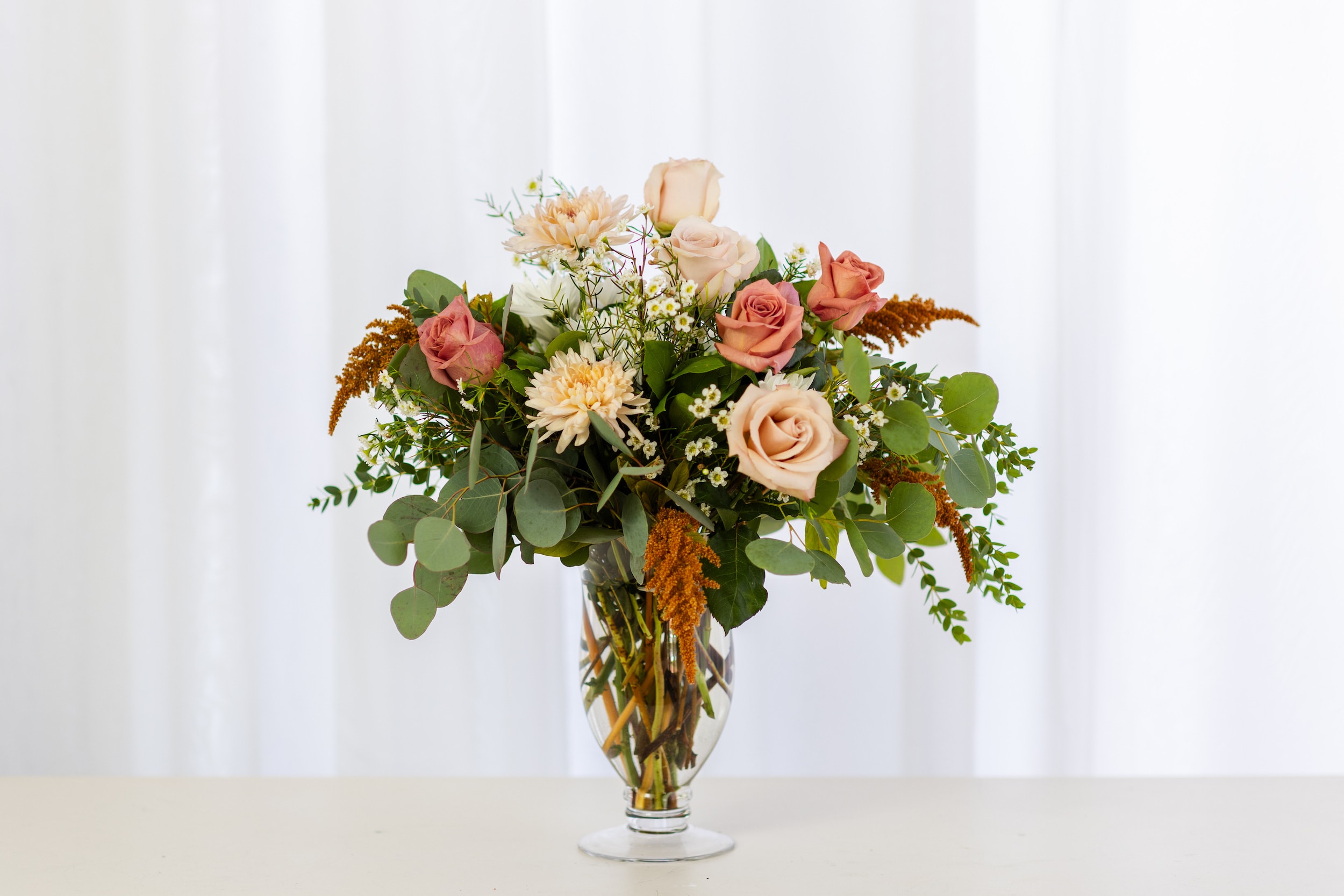 The Chai Latte - Signature Size Vase Arrangement - All the cozy vibes of warm fall spices inspired our new Chai Latte Collection. Shades of cinnamon, mocha, and cream come together in a beautiful gathering of mums, roses, berries, and branches—artfully arranged to capture the essence of the season.  Our tall arrangement is presented in an elegant glass footed vase, making it an ideal gift for an anniversary, birthday, or any special occasion.  The Chai Latte Tall Vase Arrangement measures approximately 16&quot; tall and 12&quot; wide. 
