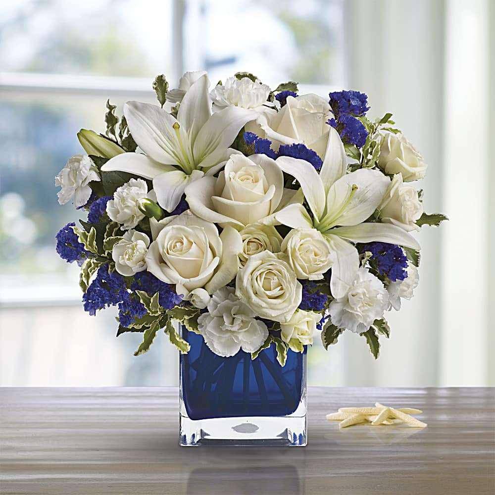 Clear Blue Skies Bouquet - Nothing better to inspire a bouquet than nature itself! Our Clear Blue Skies Bouquet is arranged to reflect the beauty of a beautiful day where the sun is shining without a cloud in the sky! Pearly white lilies surrounded by creme white roses purple statice invoke happiness in any beholder, whether they're displayed in a home or at an office, they're perfect for any kind of day. With its vibrant blue and elegant design, this bouquet is the perfect choice for those looking for exquisite flowers near me.  Includes:&nbsp; • Creme Roses&nbsp; • White Asiatic Lilies&nbsp; • White Miniature Carnations&nbsp; • Purple Statice&nbsp; • Cube Vase
