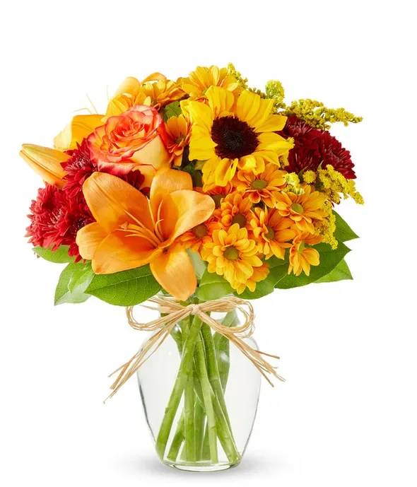 Harvest Moon Bouquet  V712325 - Capture the essence of fall with this radiant sunflower bouquet. Featuring glowing orange roses, bold orange lilies, rich maroon cushion poms, and cheerful yellow daisy poms, it's all tied together with accents of golden solidago and lush greenery. Arranged in a clear glass vase with a rustic raffia bow, this vibrant display is perfect for sending autumn cheer, celebrating special occasions, or brightening any space with seasonal beauty.