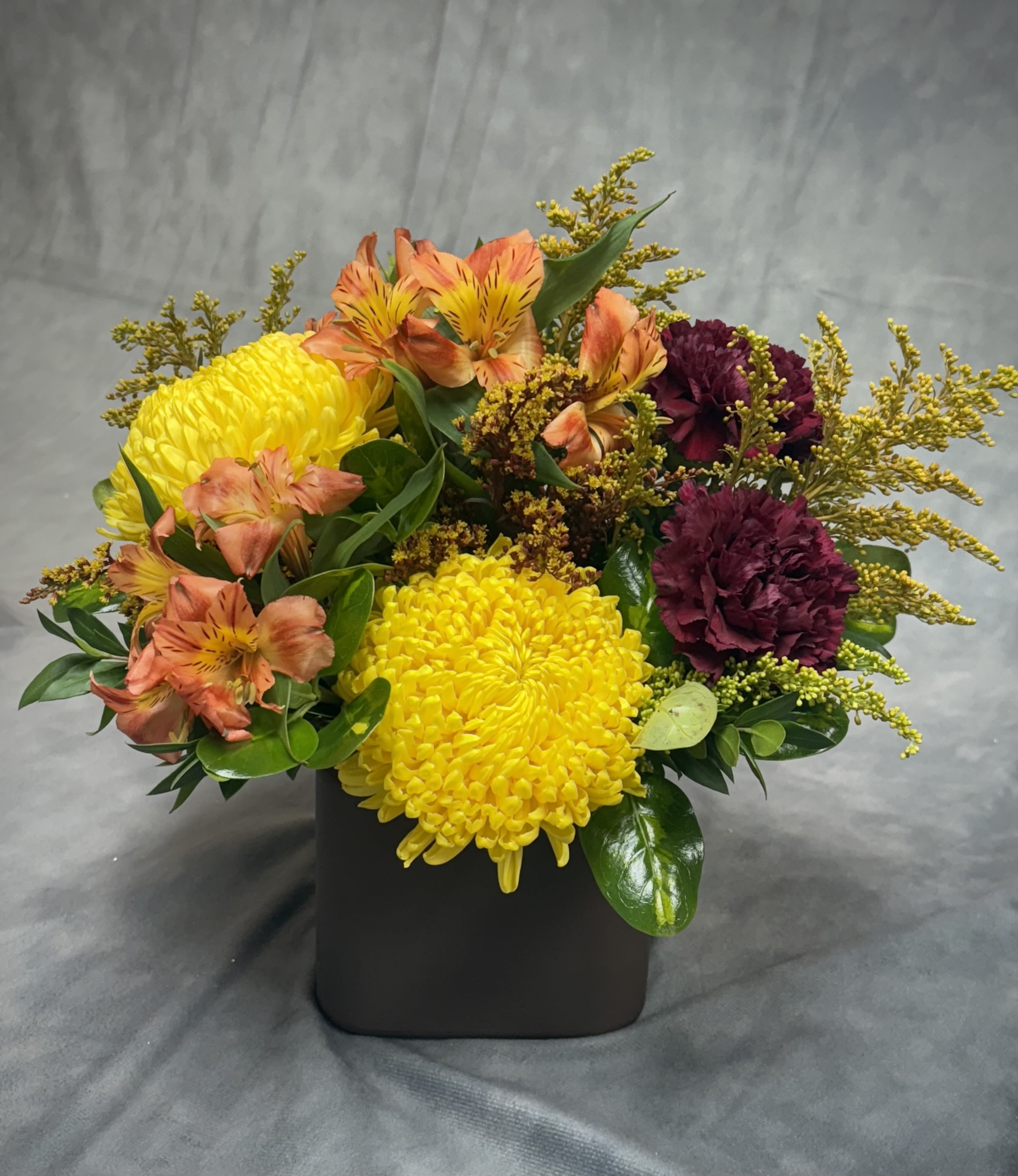 Autumn Hues Vase - A cheerful low-vase arrangement capturing the cozy colors of fall.  Featuring sunny yellow mums, rich burgundy carnations, and vibrant orange alstroemeria, this design adds a warm seasonal glow to any space — perfect for a table centerpiece or a thoughtful fall gift.
