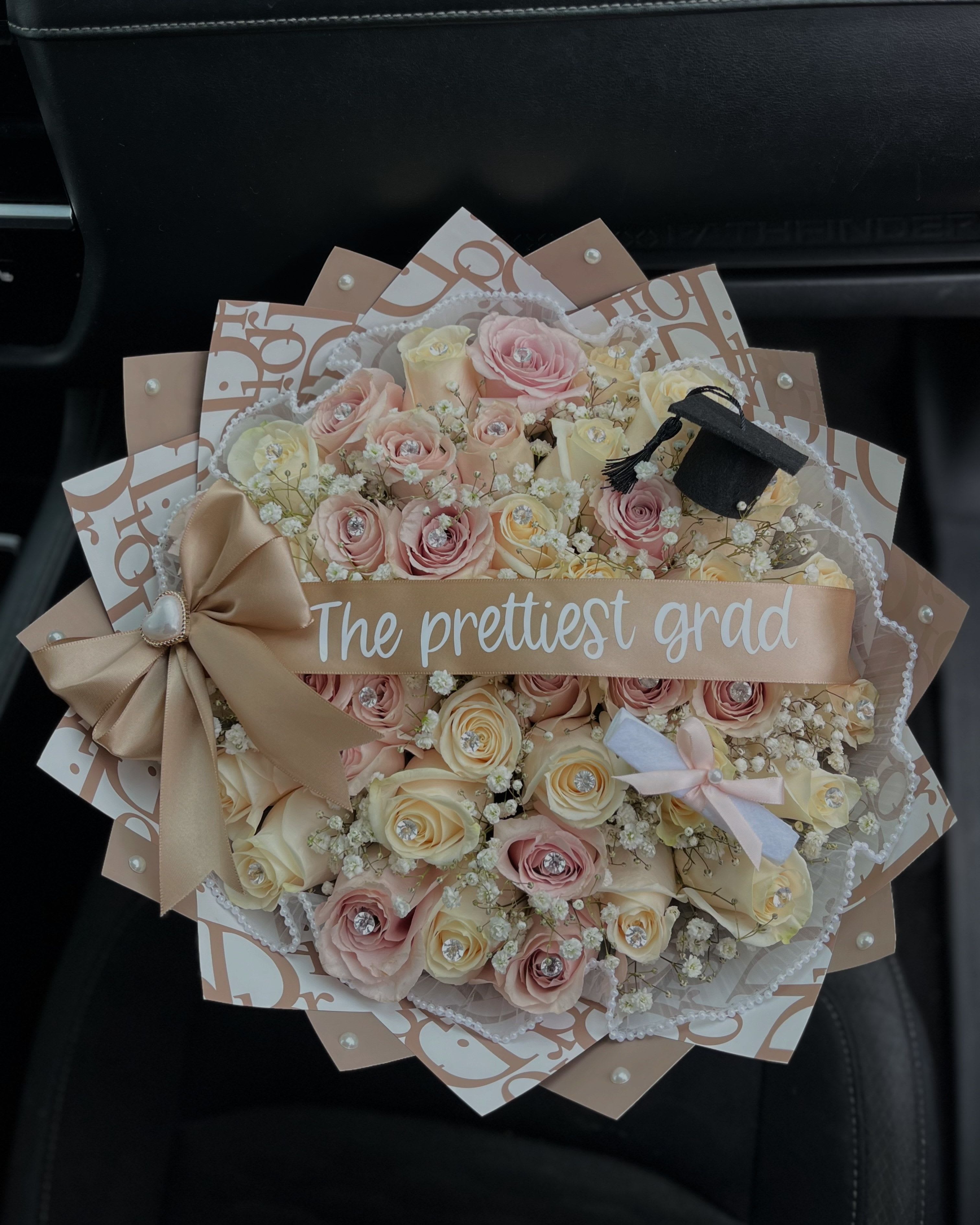 The Prettiest Grad Mauve Neutral - Beautifully balanced in soft, neutral tones of white, mocha, and mauve pink, this trendy graduation bouquet is the perfect blend of elegance and celebration. Featuring 30 white and mauve-pink roses, delicately accented with baby’s breath peeking through, it’s designed to make a stunning statement.  Each rose is adorned with a sparkling diamond pin, wrapped in premium coffee-colored designer paper adorned with pearl accents, and finished with a pearl skirt that rims the bouquet for an extra touch of class.  Topped with a mini graduation cap and diploma, and completed with a customizable ribbon and side bow, this bouquet brings a modern, stylish twist to a timeless milestone.