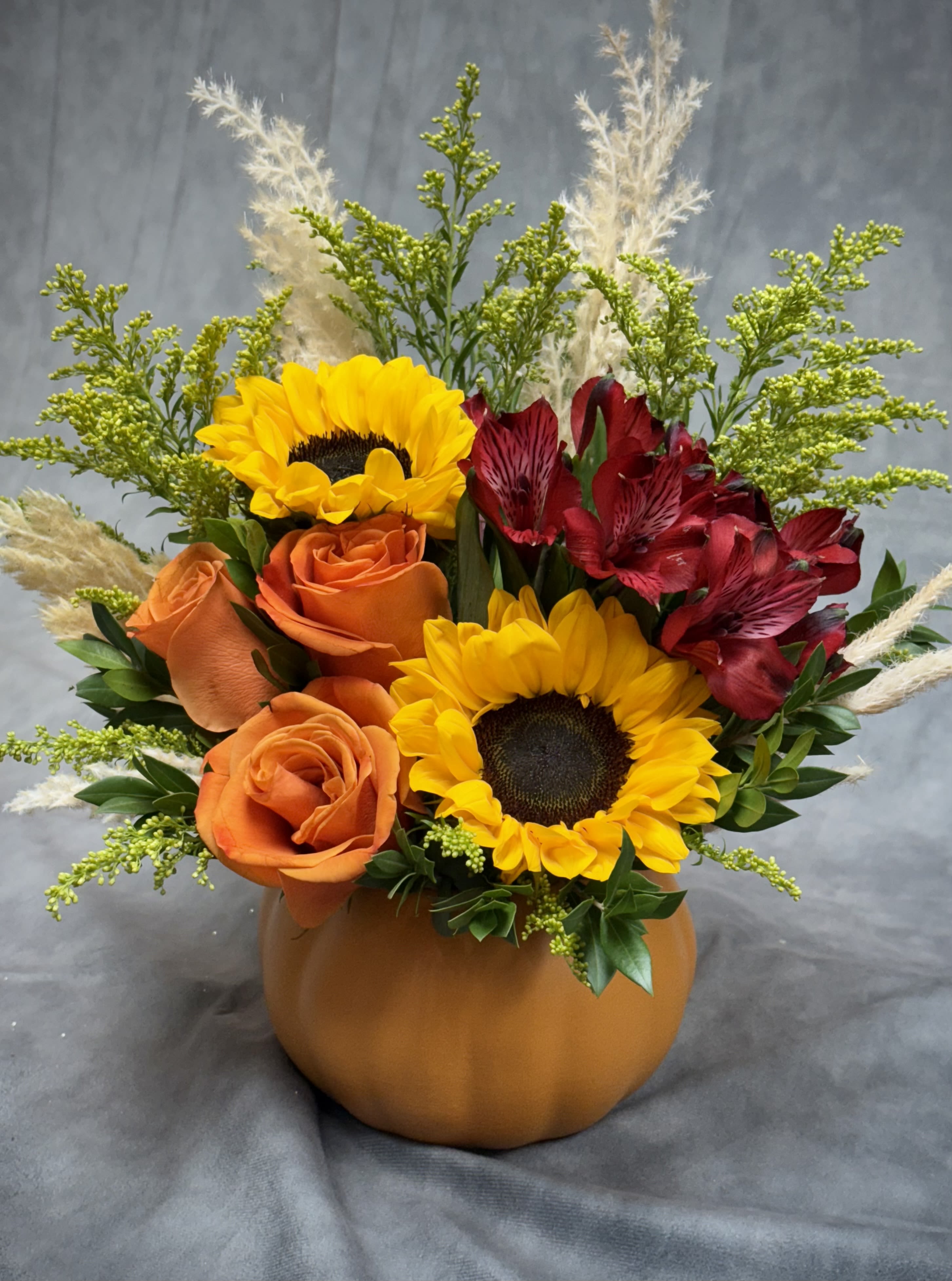 Sunny Pumpkin  - A warm, cheerful fall arrangement bursting with seasonal charm.  This design features golden sunflowers, vibrant orange roses, and rich red alstroemeria , beautifully accented with soft pampas grass inside a festive pumpkin container. Perfect for brightening any autumn table with cozy harvest vibes and sunny elegance.