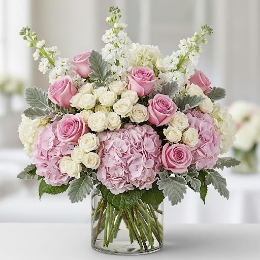 Angelina Ballerina By Black Cat Florist - Introducing Angelina Ballerina — a graceful and full floral arrangement that dances with elegance. Soft light pink roses and delicate white spray roses bloom alongside lush pink hydrangeas, while white stock flowers add airy height. Dusty miller weaves through the design with its silvery-green touch, creating a dreamy balance. Presented in a sleek clear glass cylinder vase, this arrangement captures the beauty and poise of a ballerina in bloom. 