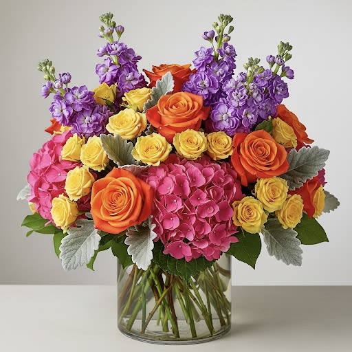 Felicia By Black Cat Florist Felicia By Black Cat Florist - Felicia — A vibrant spell of color and joy. Orange roses glow like firelight, golden yellow spray roses radiate warmth, and hot pink hydrangeas burst with bold energy, purple stock rise through the design, casting a regal, enchanting note, while dusty miller softens the palette with its silvery haze. Lush, whimsical, and full of life, Felicia is a floral celebration that feels like sunshine wrapped in mystery.