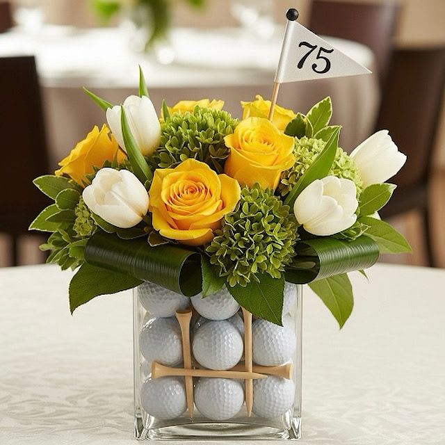 Tee-Time Bloom By Black Cat Florist  - Tee-Time Bloom Golf Centerpiece  This vibrant and playful centerpiece is a perfect tribute to the love of golf. The clear glass cube vase beautifully showcases a collection of golf balls and wooden tees, creating a unique and eye-catching base. Above, a cheerful arrangement of bright yellow roses, crisp white tulips, and lush green hydrangeas (or similar textured green blooms) bursts forth. A small flag with a customizable number – ideal for celebrating a special birthday or event – adds a personalized touch, making this a truly memorable and spirited design.