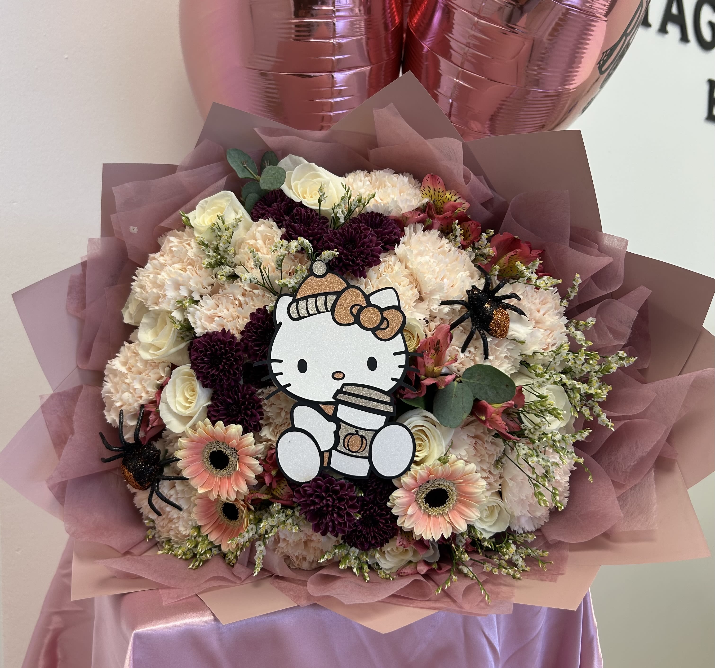 Hello Spooky Kitty Bouquet - Celebrate Halloween with a soft pastel twist and a touch of burgundy magic! This medium-sized bouquet combines white roses, soft blush-salmon carnations, gerberas, and rich burgundy blooms, accented with delicate filler flowers and wrapped in blush pink paper. Featuring an adorable Hello Kitty holding a coffee cup, and two little spider accents, this bouquet blends cute charm and spooky vibes in one perfect design — the sweetest way to say &quot;for my Boo, with love.&quot; + Limited seasonal design - available only during October!