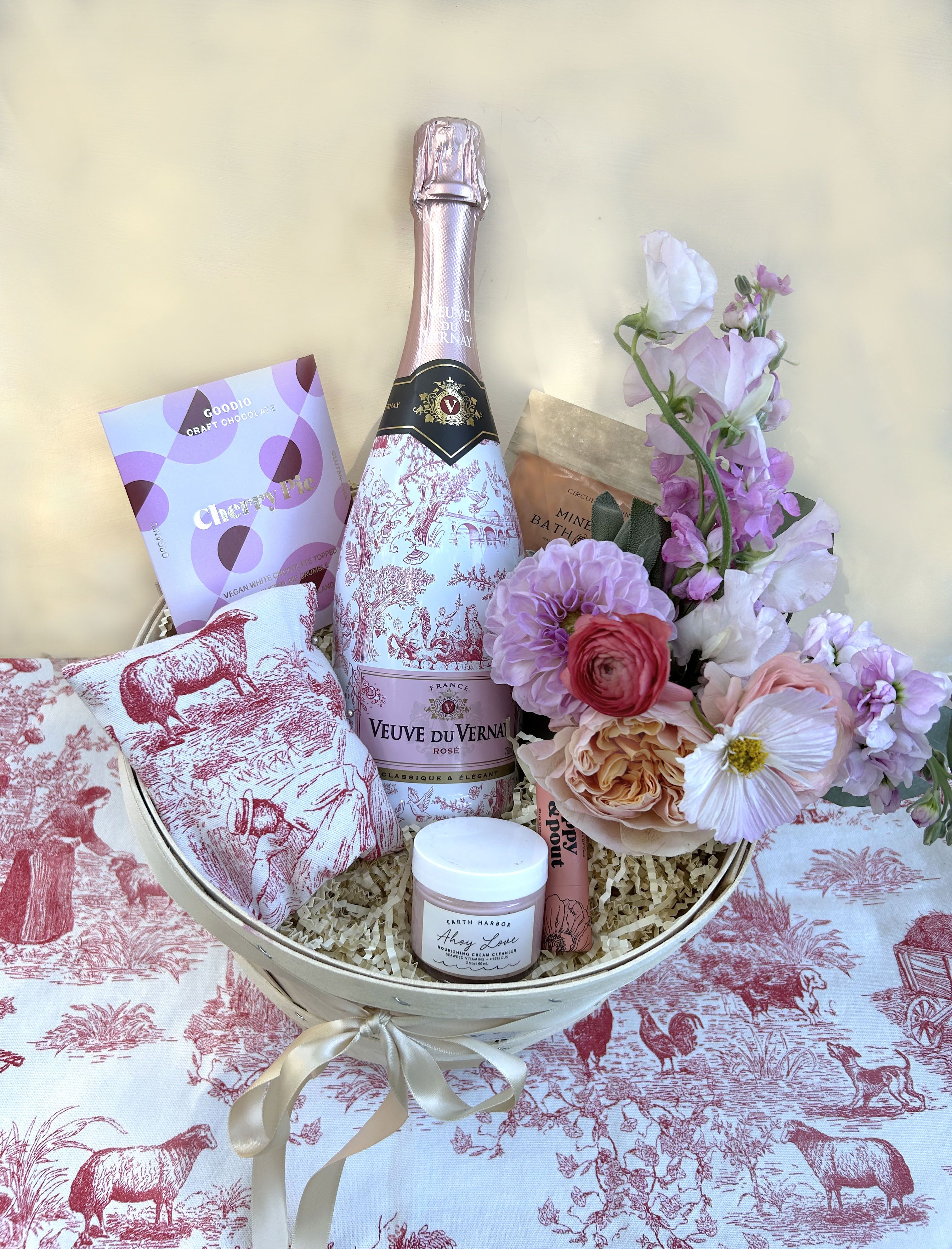 Toile de Jouy Basket - Classic and elegant basket in the theme of the historic fabric from Jouy, France. The beautiful Pastoral romantic pattern is world famous. Here it's featured on the Veuve du Vernay Brut Rosé Limited Edition bottle of French bubbles and in the lavender sachet made in the Toile de Jouy fabric. The European Cherry Pie chocolate (V and GF) compliments the feeling of this extravagant basket containing wellness treats. Seaweed and Hibiscus vitamin facial creme, Poppy and Pout Pink Grapefruit lip balm. A lip balm formula as smooth as can be. Each cardboard tube is filled from the bottom to the top with lip hydrating goodness. Circular Living Rose petal and Geranium bath soak: A luxurious mineral soak for sore muscles with rose, geranium, and lemon to balance the skin and cleanse the mind and body. Mini Blomst arrangement placed in a small clear cyllinder vase. 