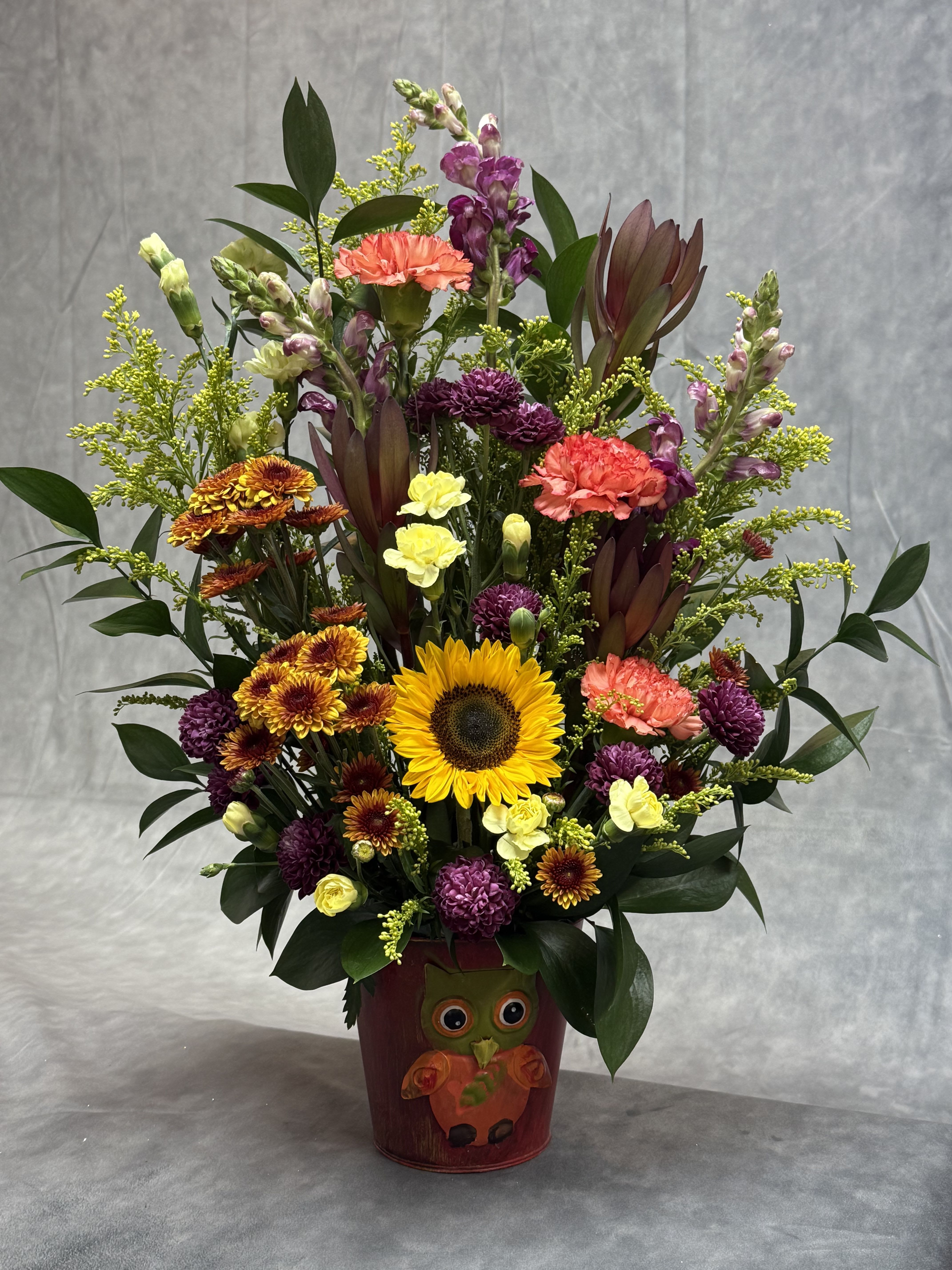 Fall Hoot &amp; Bloom  - Celebrate the cozy charm of autumn with a whimsical mix of sunflowers, orange and purple blooms, and rustic textures, all gathered in a festive owl container that brings fall cheer to any space.