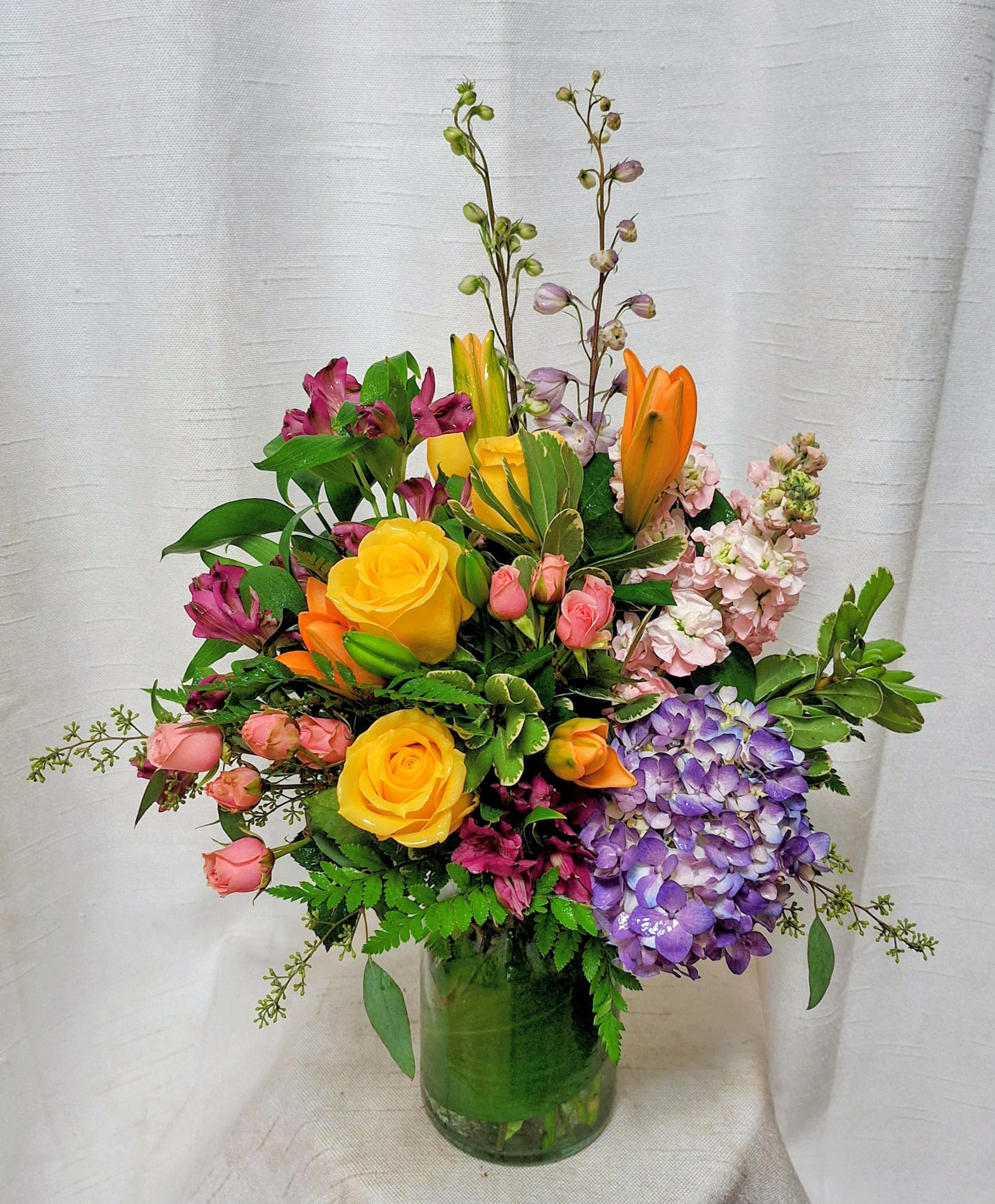 Happy Days (DL) - Elevate your space with our modern floral arrangement, thoughtfully crafted in a clear glass leaf-lined vase. This artistic ensemble features a vibrant mix of orange lilies, delphinium, stock, spray roses, purple hydrangea, yellow roses, and assorted greenery. 