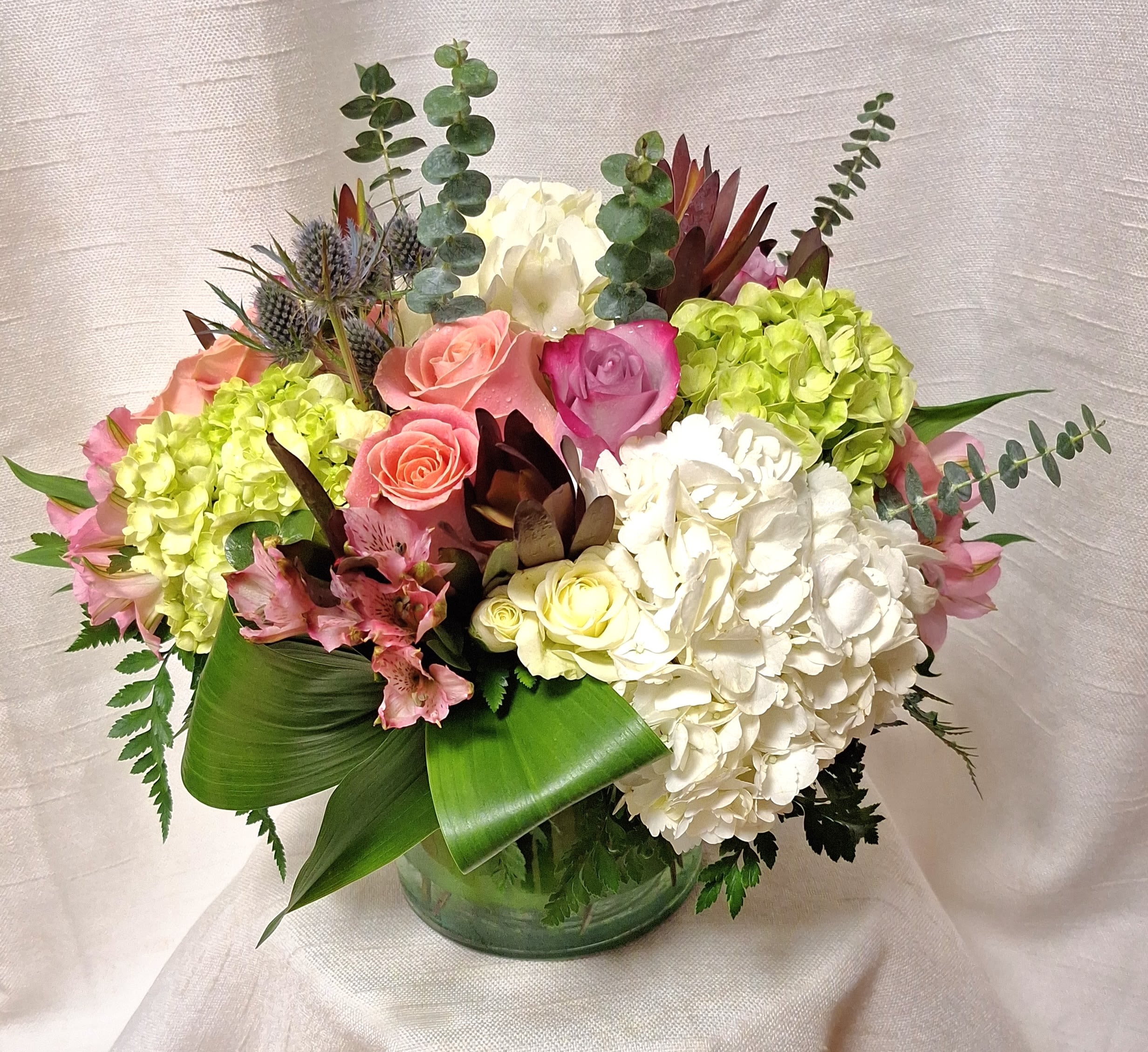 Floral Mosaic (DL) - Add a contemporary touch to your décor with our modern and colorful floral arrangement in a clear 6x6&quot; glass cylinder vase. This stunning ensemble combines the softness of green and white hydrangea, the vibrancy of pink and lavender roses, and the delicate charm of gum thistle and more for a sophisticated and stylish look. Perfect for any occasion, this arrangement is sure to impress.  