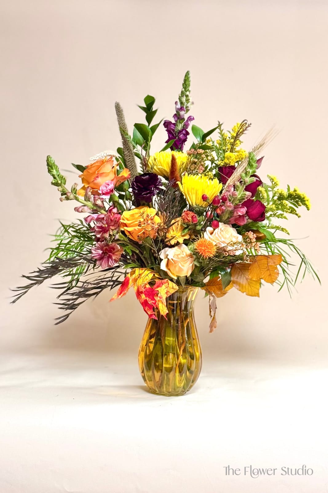 Autumn Sunburst - This stunning fall-inspired design bursts with the rich, warm colors of the season. Featuring vibrant orange and peach roses, golden spider mums, burgundy blooms, and touches of yellow solidago, the arrangement is beautifully accented with seasonal foliage, textural grasses, and autumn leaves. Designed in an amber glass vase, it brings the perfect blend of rustic charm and elegance—ideal for brightening your home or gifting during the fall season.
