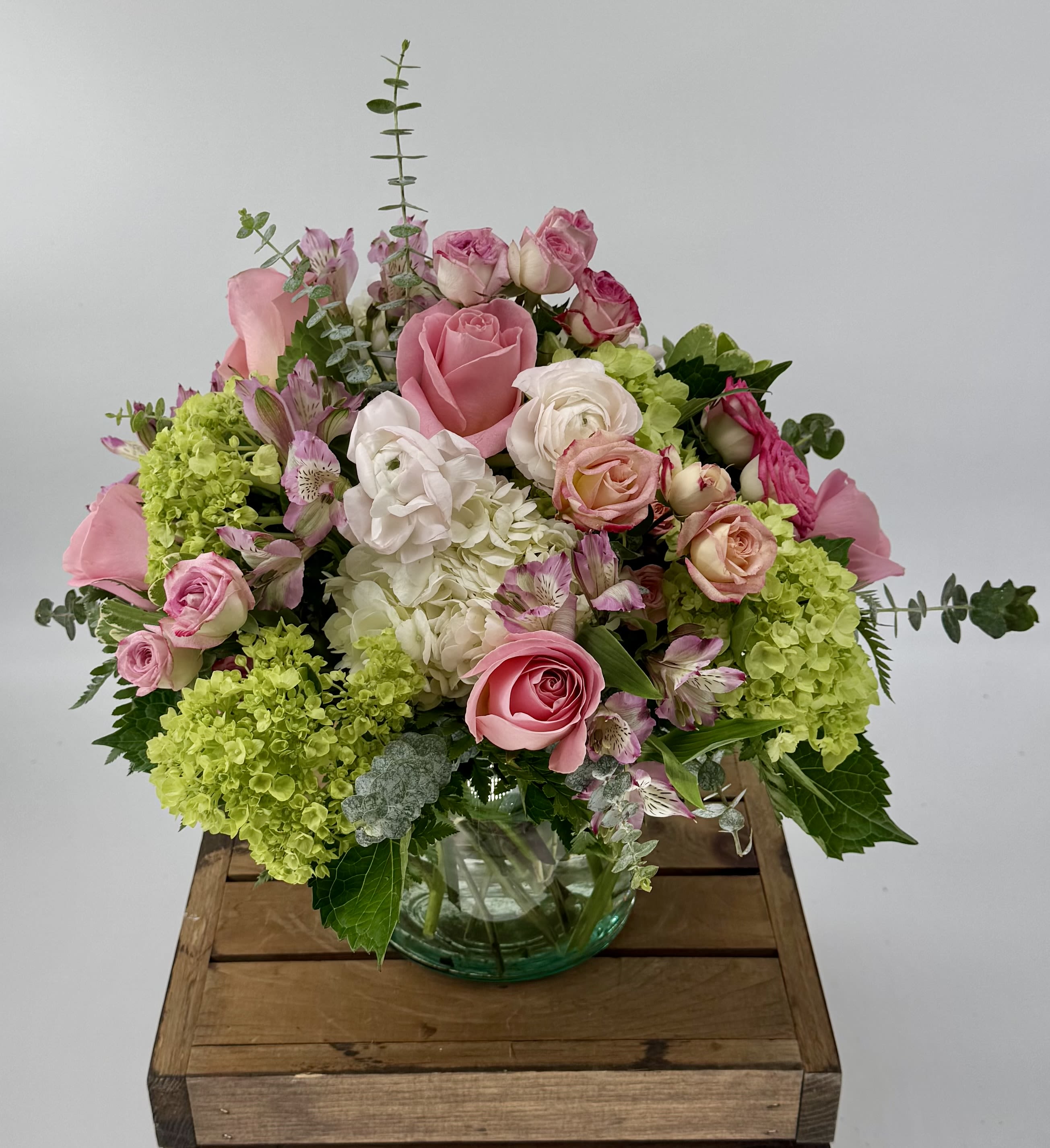 Simplicity (DL) - Delight in a stunning blend of green and white hydrangea, pink ranunculus, roses, alstroemeria, and eucalyptus, artfully arranged in a leaf-lined 6x6” clear vase—crafted to elevate your most cherished moments. 
