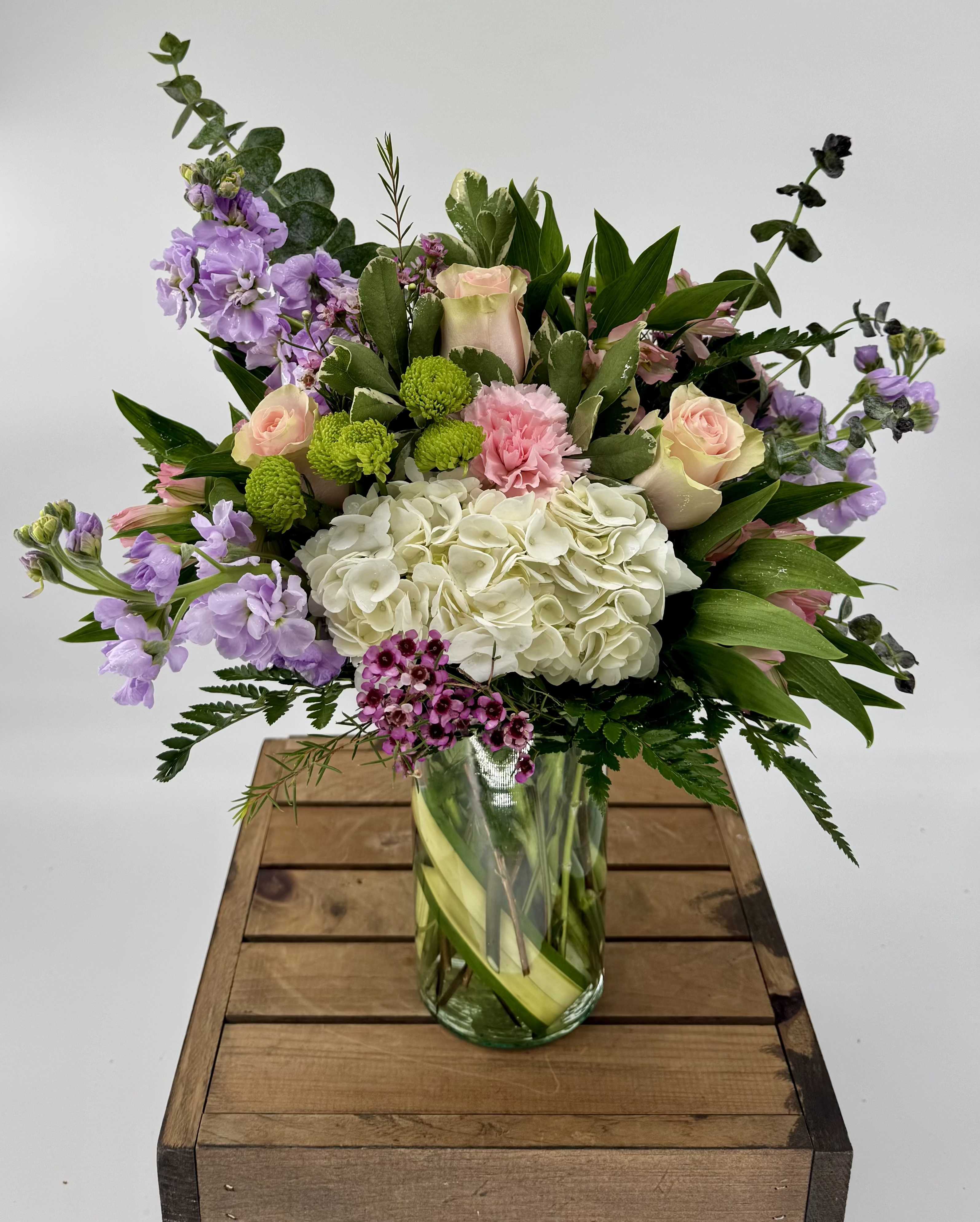 The April (DL) - This enchanting bouquet features an exquisite combination of pink roses, white hydrangeas, lavender stock, white carnations, pink alstroemeria, green button poms, and lush greenery, perfectly arranged in a traditional glass vase.