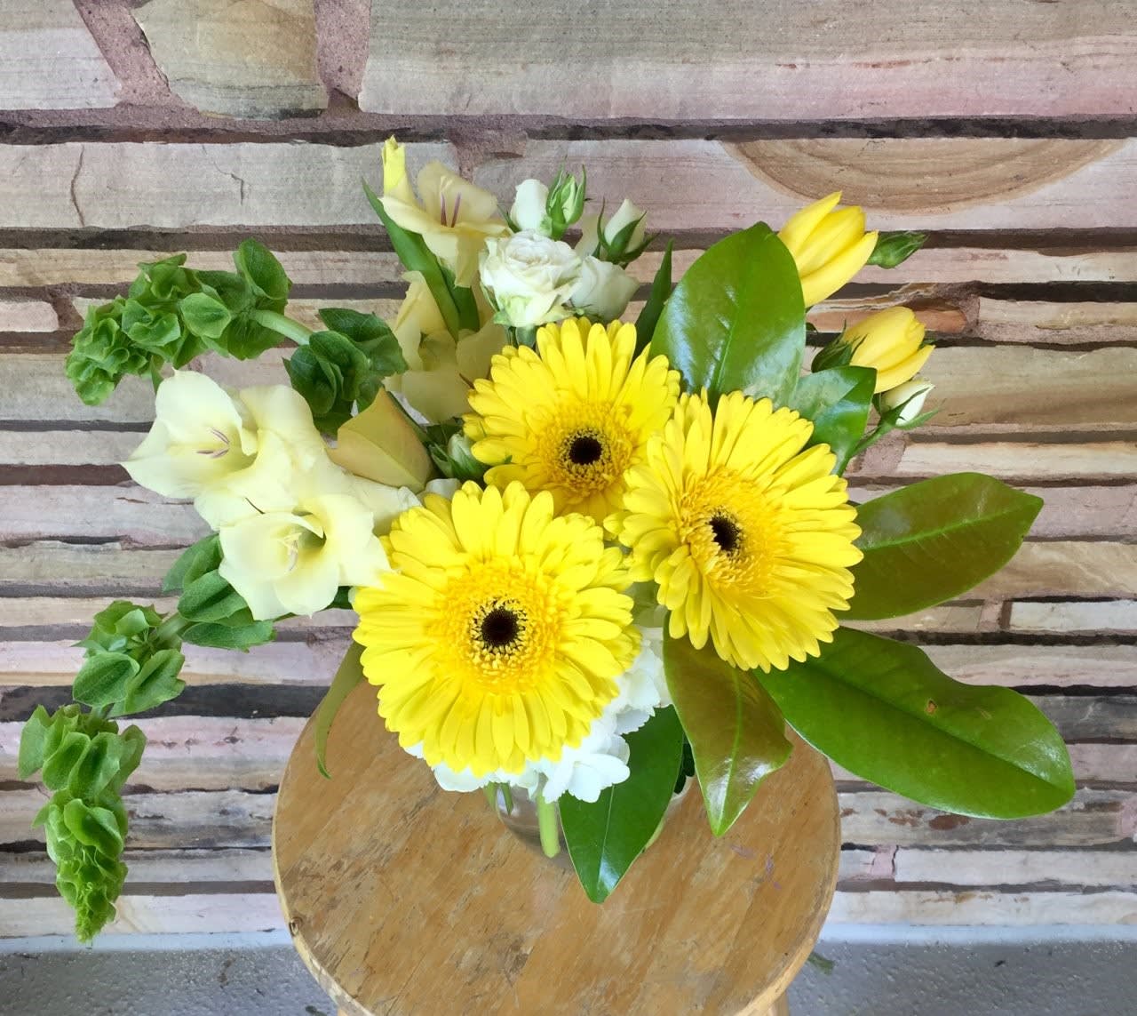 The Sun of Africa The Sun of Africa - Yellow gerbs yellow tulips white spray roses bells of Ireland hydrangea magnolia leaves glads with glass vase and other assorted greens.