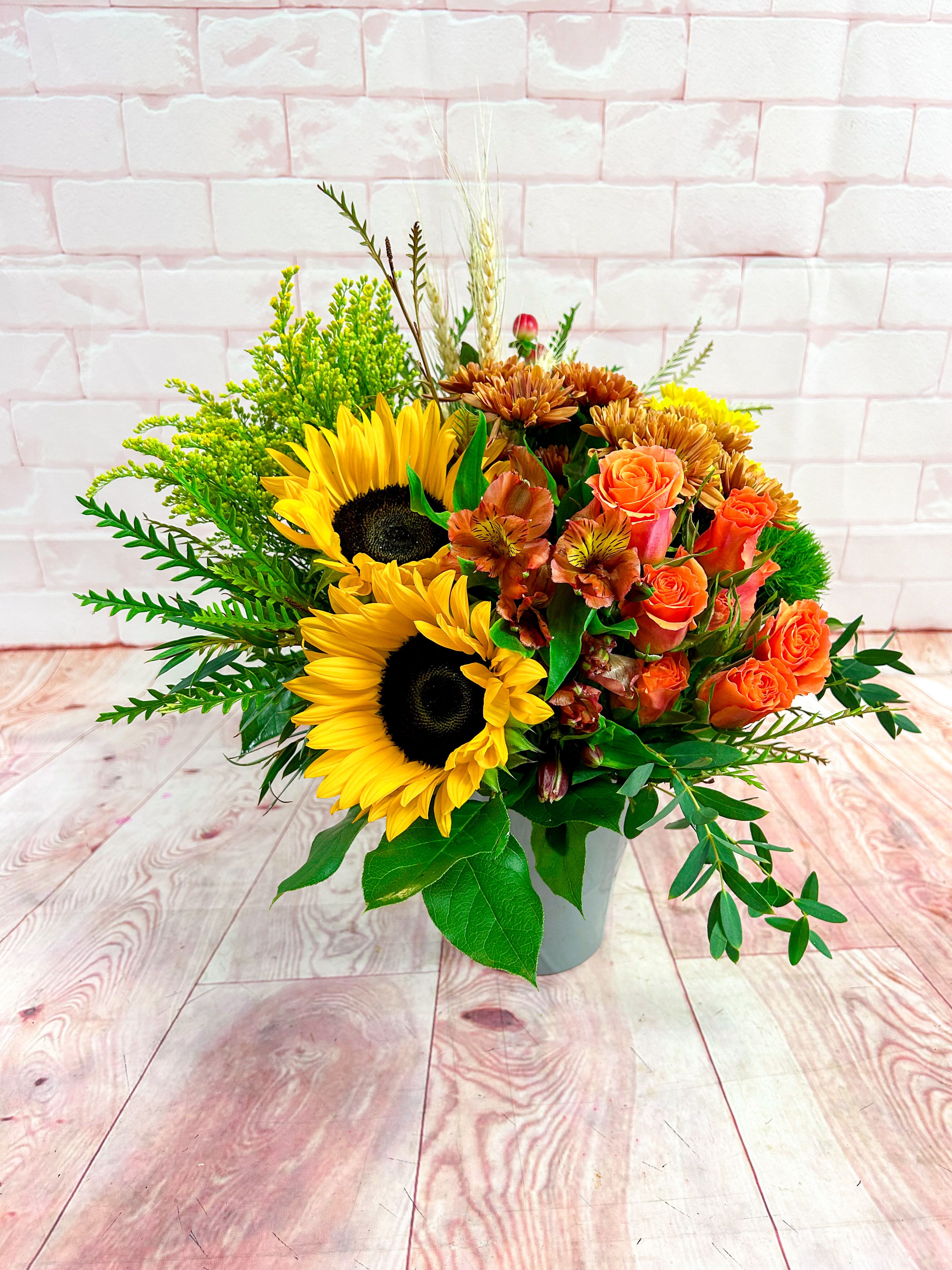 Sunflower Spice Bouquet - Bring the golden glow of fall into any space with our Harvest Sunshine Bouquet. Bursting with radiant sunflowers, vibrant orange spray roses, rust-colored mums, and seasonal accents of wheat and greenery, this cheerful design perfectly captures the cozy beauty of autumn days.  Arranged in a modern ceramic vase, it’s an inviting gift for Thanksgiving, birthdays, or simply to brighten someone’s day. Its rich textures and sunset tones make it a stunning seasonal centerpiece or a thoughtful gesture of warmth and gratitude.