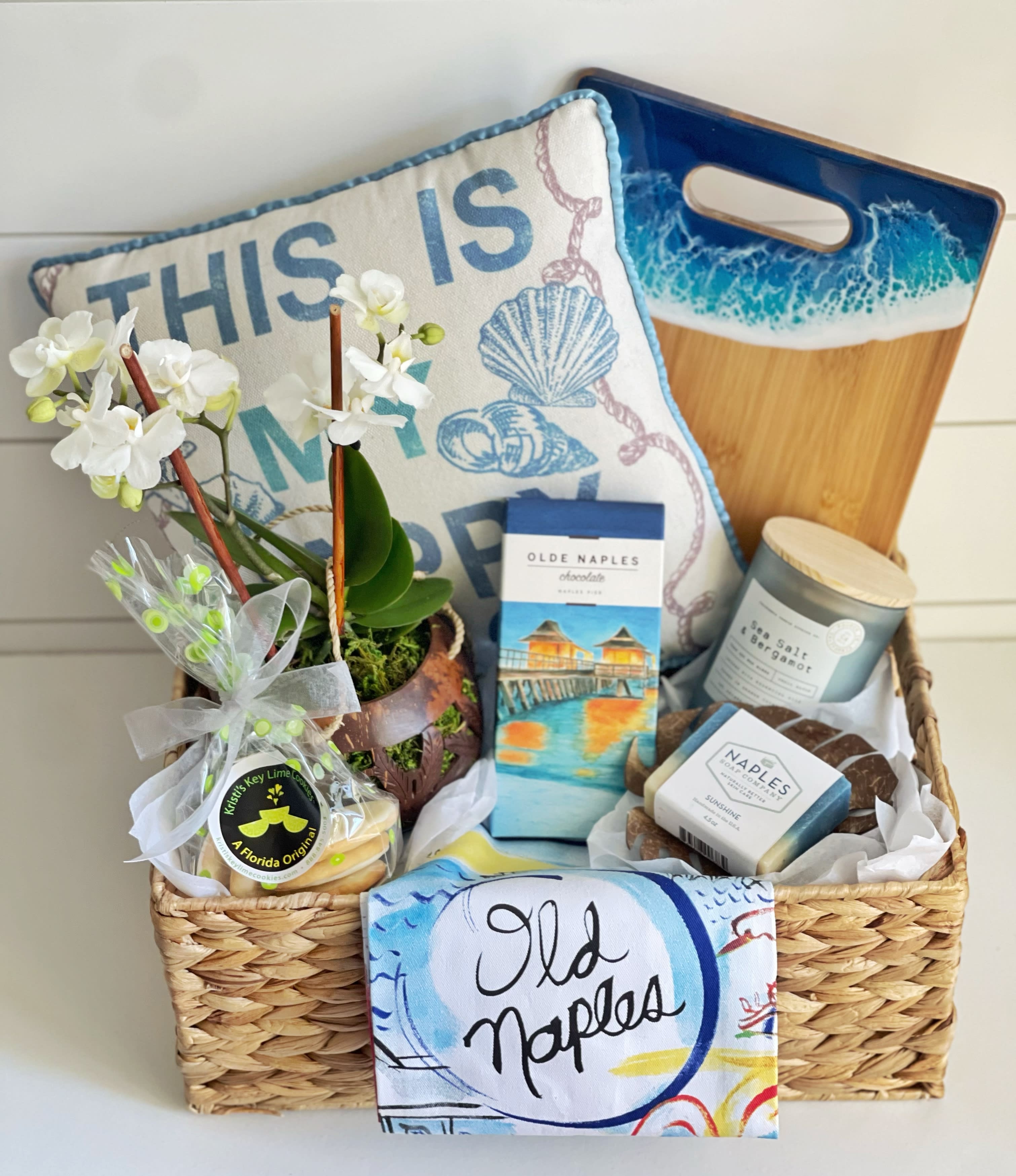 Coastal Gift Basket - This basket really captures the feel of living by the ocean. It includes: -hand/ tea towel with original art of popular Naples locations  -THIS IS MY HAPPY PLACE decorative pillow  -Naples Soap Company, bar soap with soap dish -Coastal cutting board/serving board/cacciatore board, made from Florida artist - sea glass candle with ocean scent - Beautiful Live Orchid in a unique coconut plant hanger  -Kristi’s Key Lime Cookies, 1/4 lb. bag, vanilla cookies smothered with decadent key lime topping… totally addicting! Kristi’s Key Lime Cookies were featured in the Daytime EMMY® Awards Swag Bags in Hollywood CA and the News &amp; Documentary Emmy® Award Swag Bags in New York City. -Organic dark chocolate with sea salt bar from Olde Naples Chocolates (box cover is of Naples pier) Quality reusable Basket 