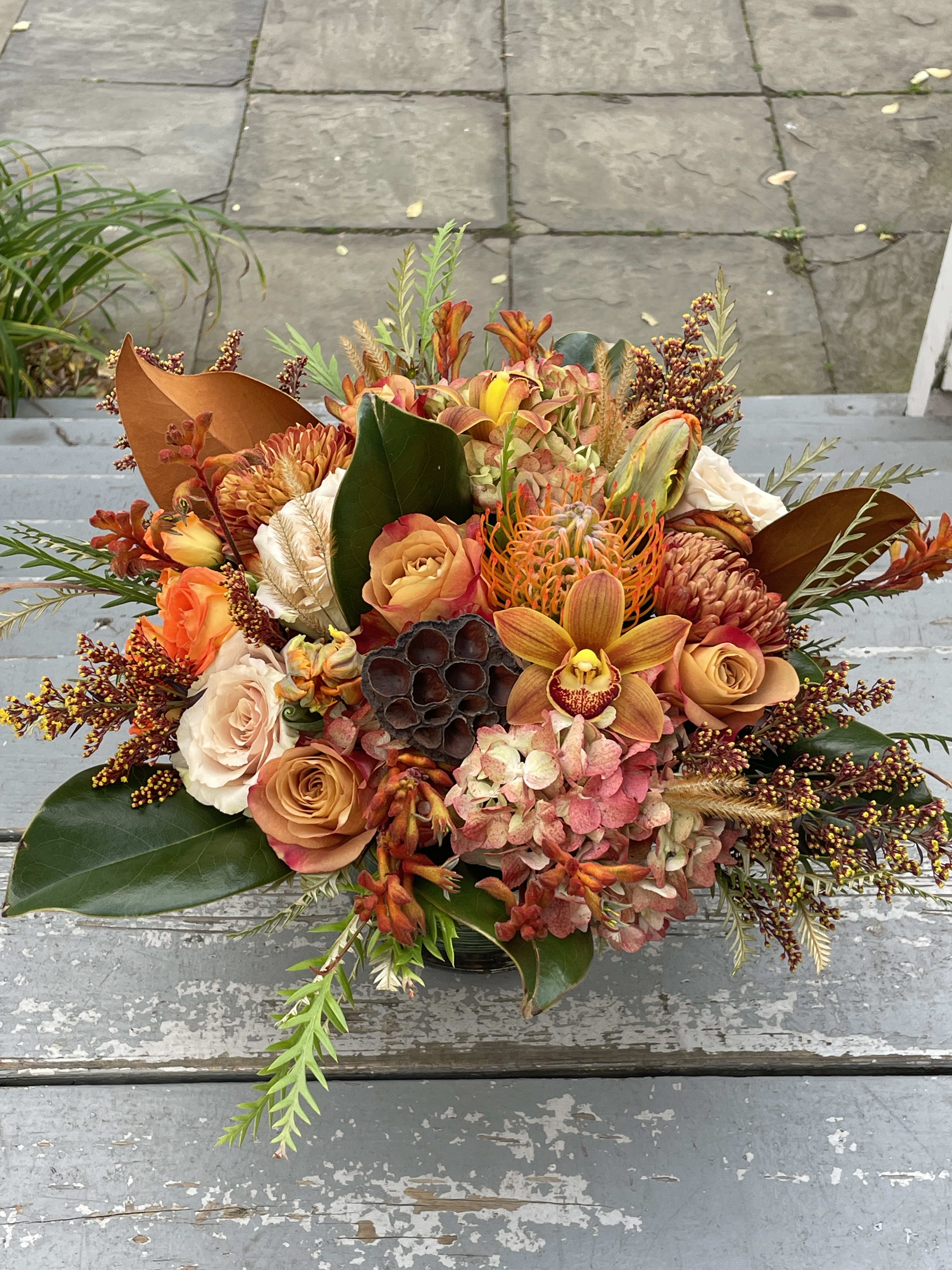Give Thanks - A low, lush centerpiece for your holiday table. Featuring the best colors and textures of the season. Arrangements may vary but you can expect to find floral varieties such as cymbidium orchids, roses,  mums, pincushion protea, kangaroo paw, and lotus pods accented with beautiful fall foliage such as mangolia leaves and gravillia. Shades of rust, tan, orange, burgundy and ochre.