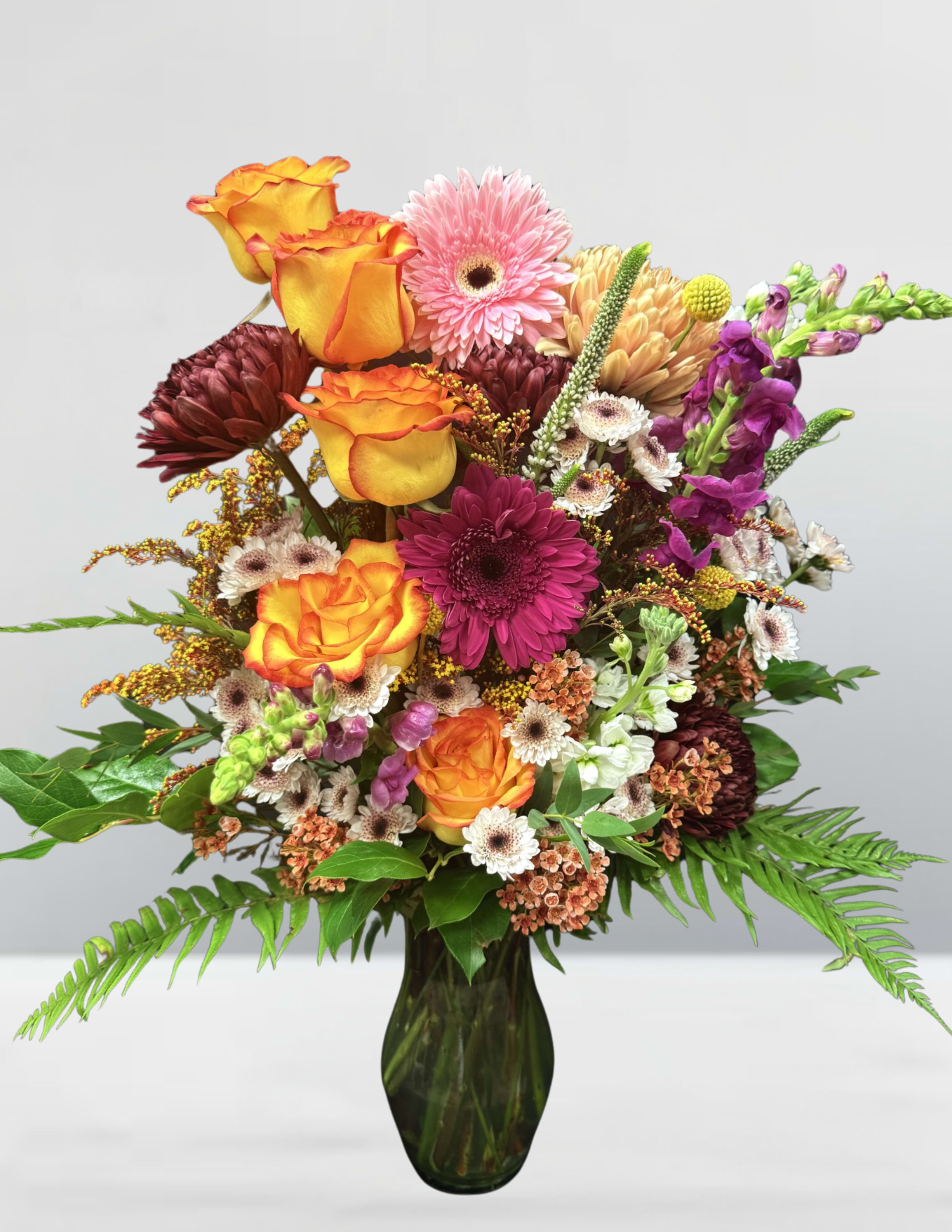 Amber & Amethyst Amber & Amethyst - Amber & Amethyst A luxurious symphony of warmth and elegance, Amber & Amethyst captures the perfect balance between fiery golden hues and deep, regal purples. Bursting with the golden glow of flame-tipped roses and accented by rich burgundy chrysanthemums and vibrant magenta gerberas, this arrangement evokes the warmth of a late autumn sunset. Delicate pink and blush gerbera daisies soften the boldness, while textured elements like snapdragons, craspedia, and sprigs of greenery add dimension and movement. White and soft peach filler blooms bring lightness to the composition, creating a harmonious contrast that makes every flower shine. Designed in a deep green glass vase to ground the palette, this bouquet is a celebration of bold beauty and timeless charm—perfect for making a statement, sharing heartfelt emotions, or simply brightening a room with elegance.