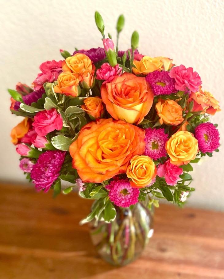 Colors of Summer  - This vibrant arrangement is definitely an eye catcher. This design includes Orange Roses, Orange Spray Roses, Hot Pink Matsumoto Asters and Mini Carnations. All set in variegated foliage and in a clear glass vase. This design measures approx. 11&quot; W x 14&quot; H.