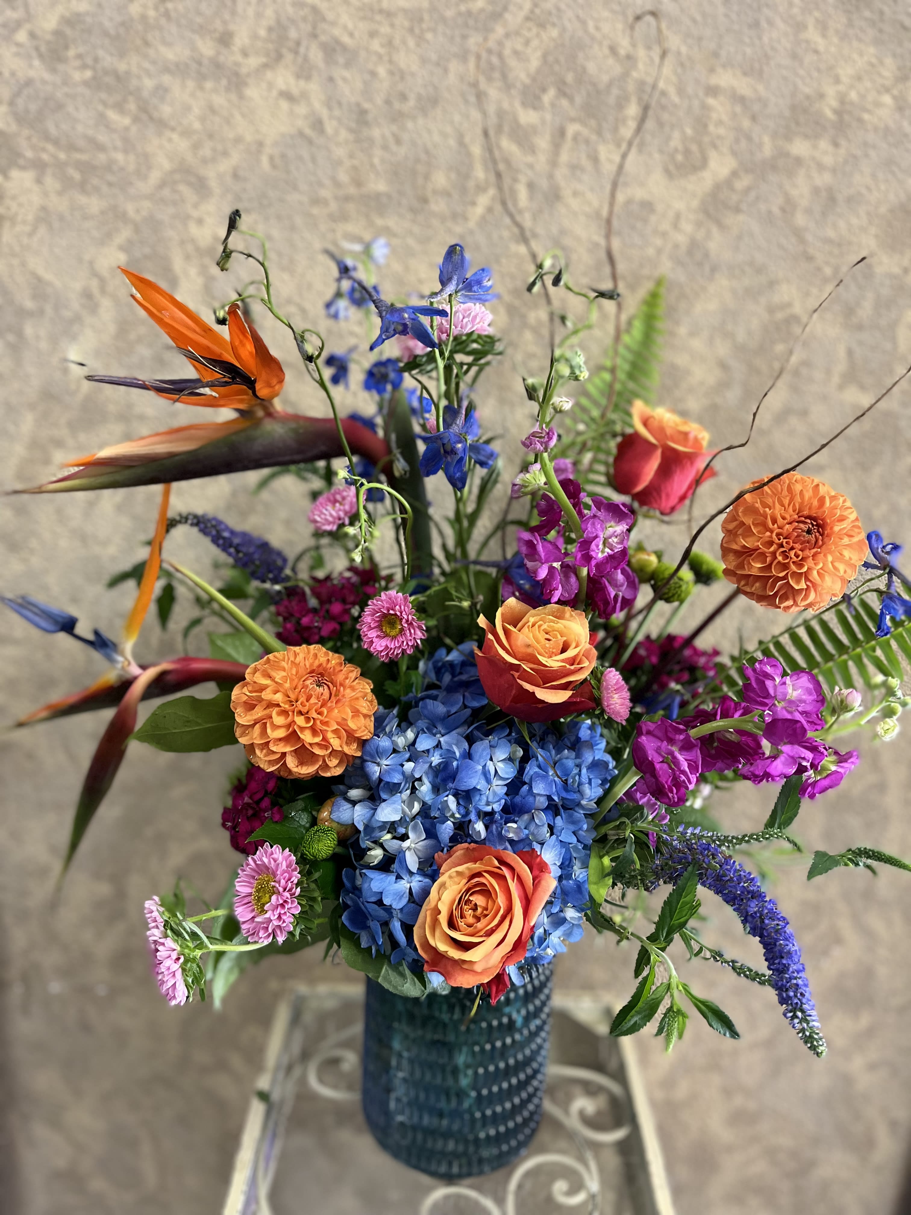Bohemian Babe Bohemian Babe - Recipe: Orange Hybrid Roses, Birds of Paradise Blue Hydrangea, Spray Roses, Larkspur, Orange Dahlias, Purple Stock, Pink Matsumoto, and more in a blue glass vase. Season: Seasonal Dahlias (mid-summer to November) Design View: Front Facing View Photo shown: Standard