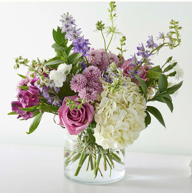 Lavender Twist Lavender Twist - A whimsical dance of soft purples, creamy whites, and lush greenery, this arrangement is pure floral joy in a vase. Blush-toned roses, playful lavender blooms and white hydrangeas. Artfully gathered in a clear glass cylinder, this bouquet brings a sense of effortless beauty!