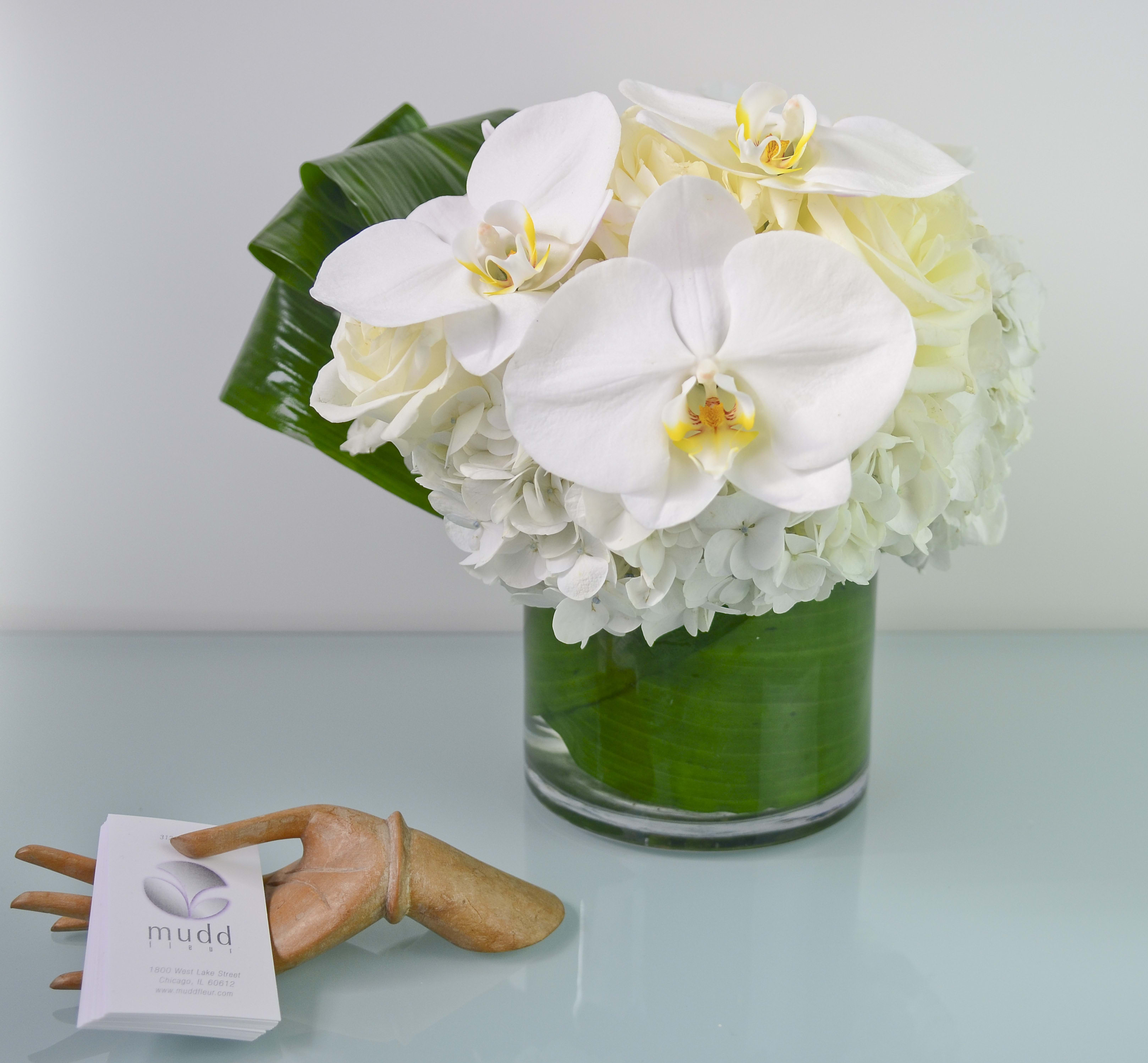 Pure White Perfection - White orchid blooms, roses, hydrangea and rolled ti-leaves styled in a clear glass vase lined with a green leaf wrap. 