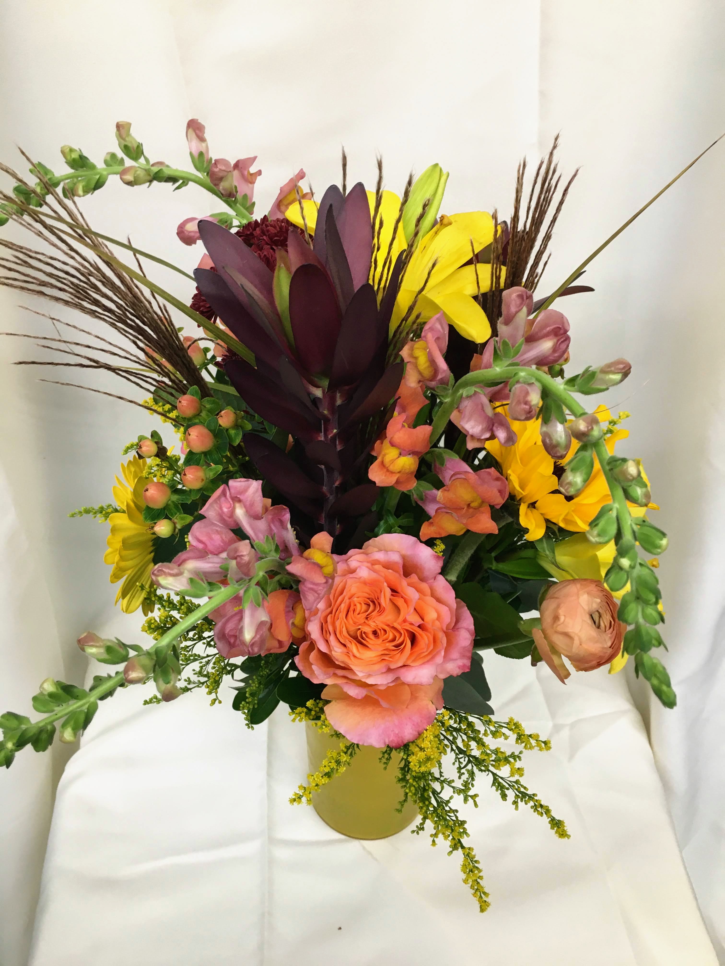 Autumnal Textures Autumnal Textures - Safari Leucadendron, peach ranunculus, roses, lilies, snapdragons, sunflowers, solidago, and ornamental grasses arranged into a taller colored glass vase.