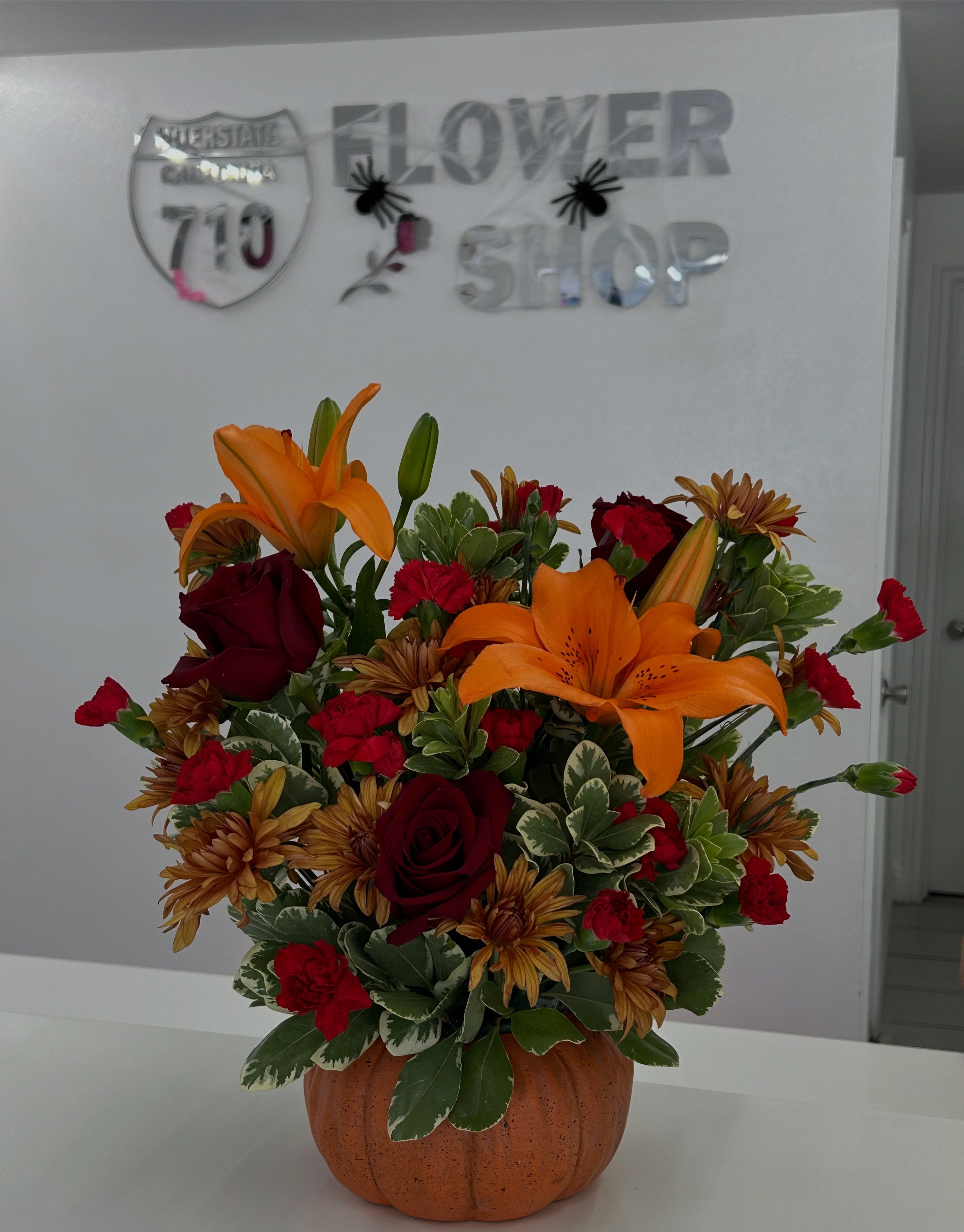 Pumpkin Spice Bloom - Bring the cozy feeling of fall indoors with this festive mix of orange lilies, red roses, and golden cushion pompoms arranged in a pumpkin vase! (If some flowers and/or the vase are not available there will be minor substitutions.)