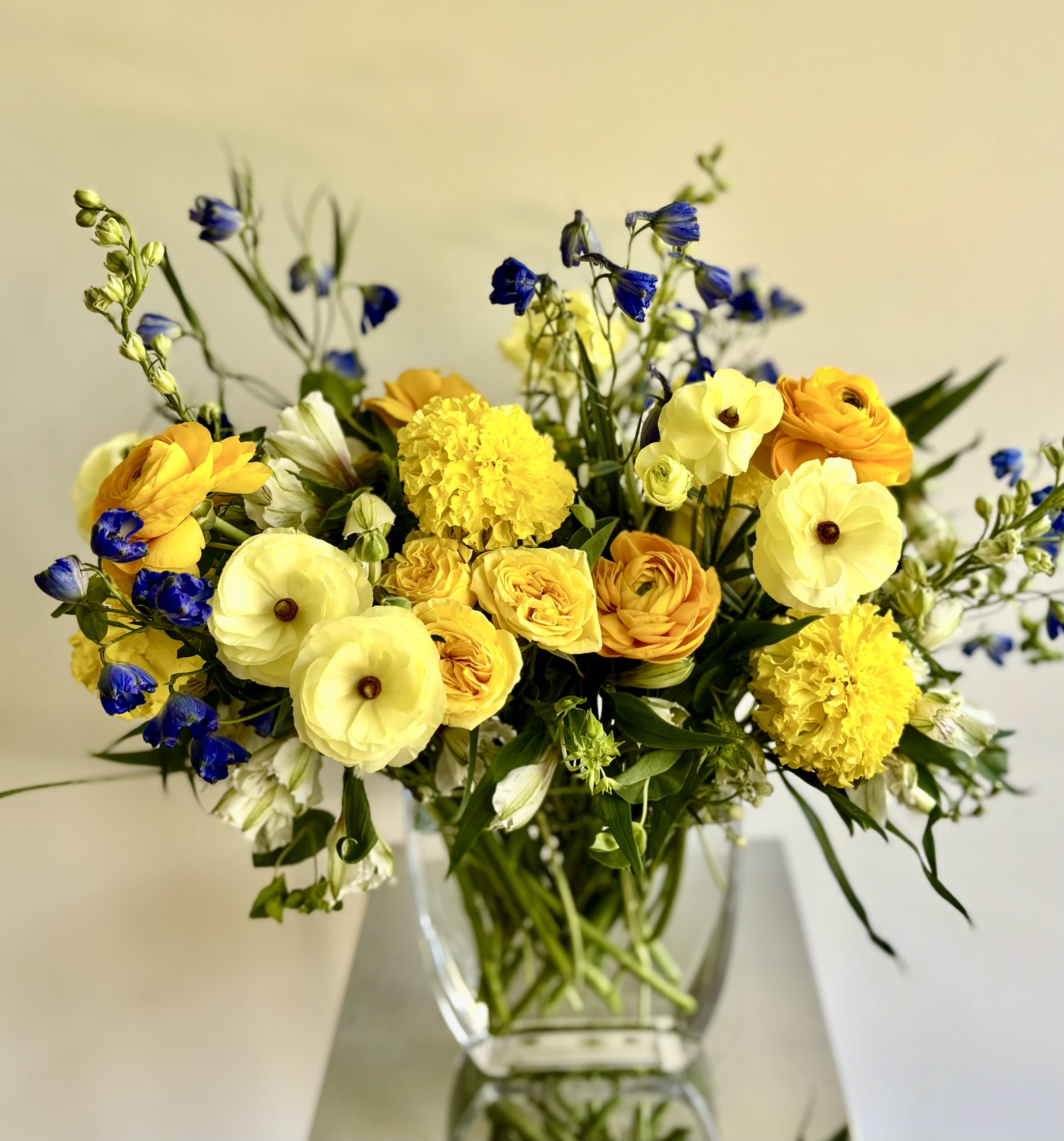 Melinda - Lovely garden-style flower arrangement with blue-yellow-white blooms in a glass vase. Michaela carefully selects the freshest flowers with the mix of roses, lisianthus, delphinum, ranunculus, tulips when available, bupleurum and other premium flowers and fillers to make this flower arrangement sunny and happy.