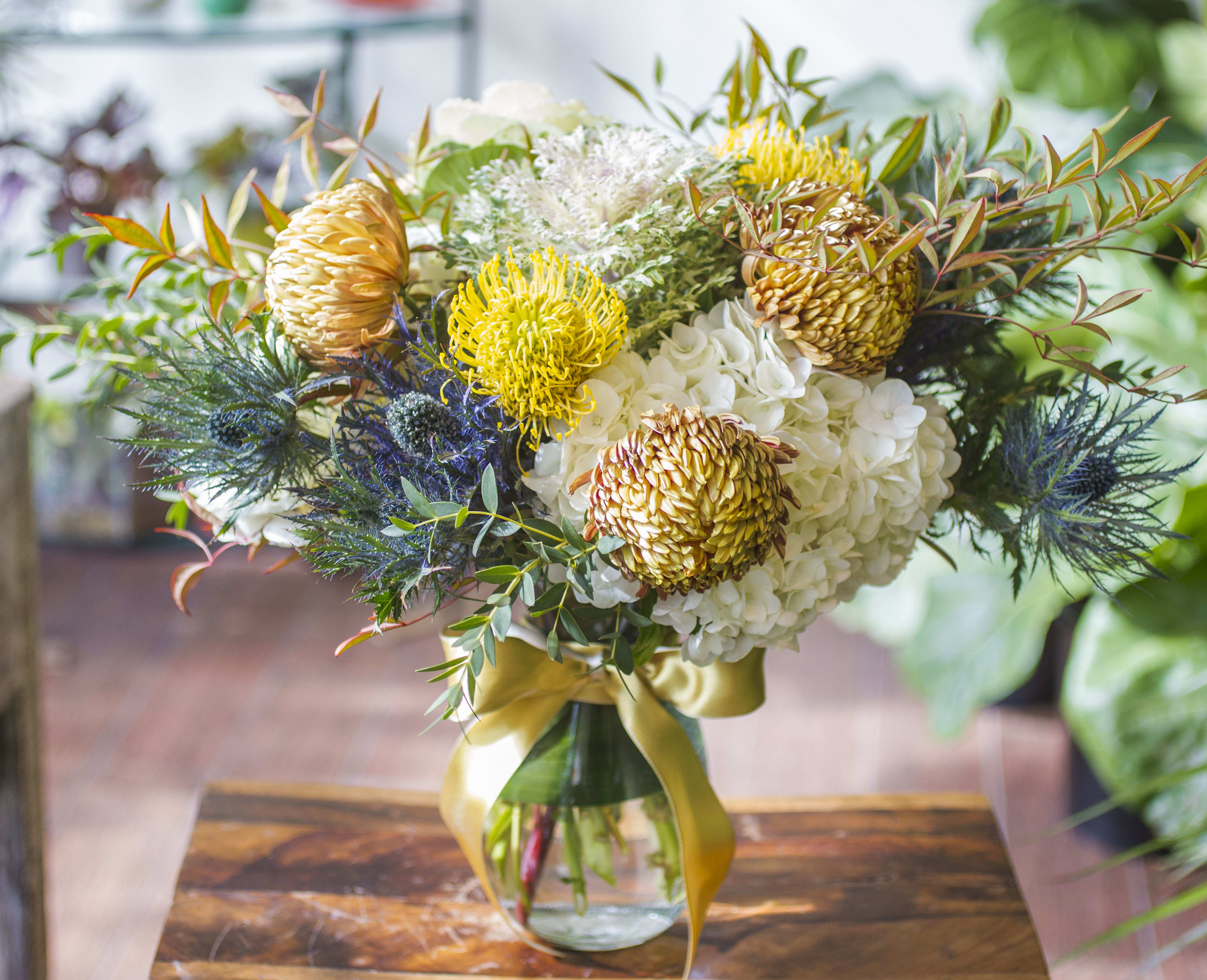 Isabella - A lovely, gentle blend of warm and cool tones, perfect for autumn. Pin-Cushion Protea, Chrysanthemum, Thistle, Kale and Hydrangea with autumn foliage, arranged in a clear glass vase with ribbon. 