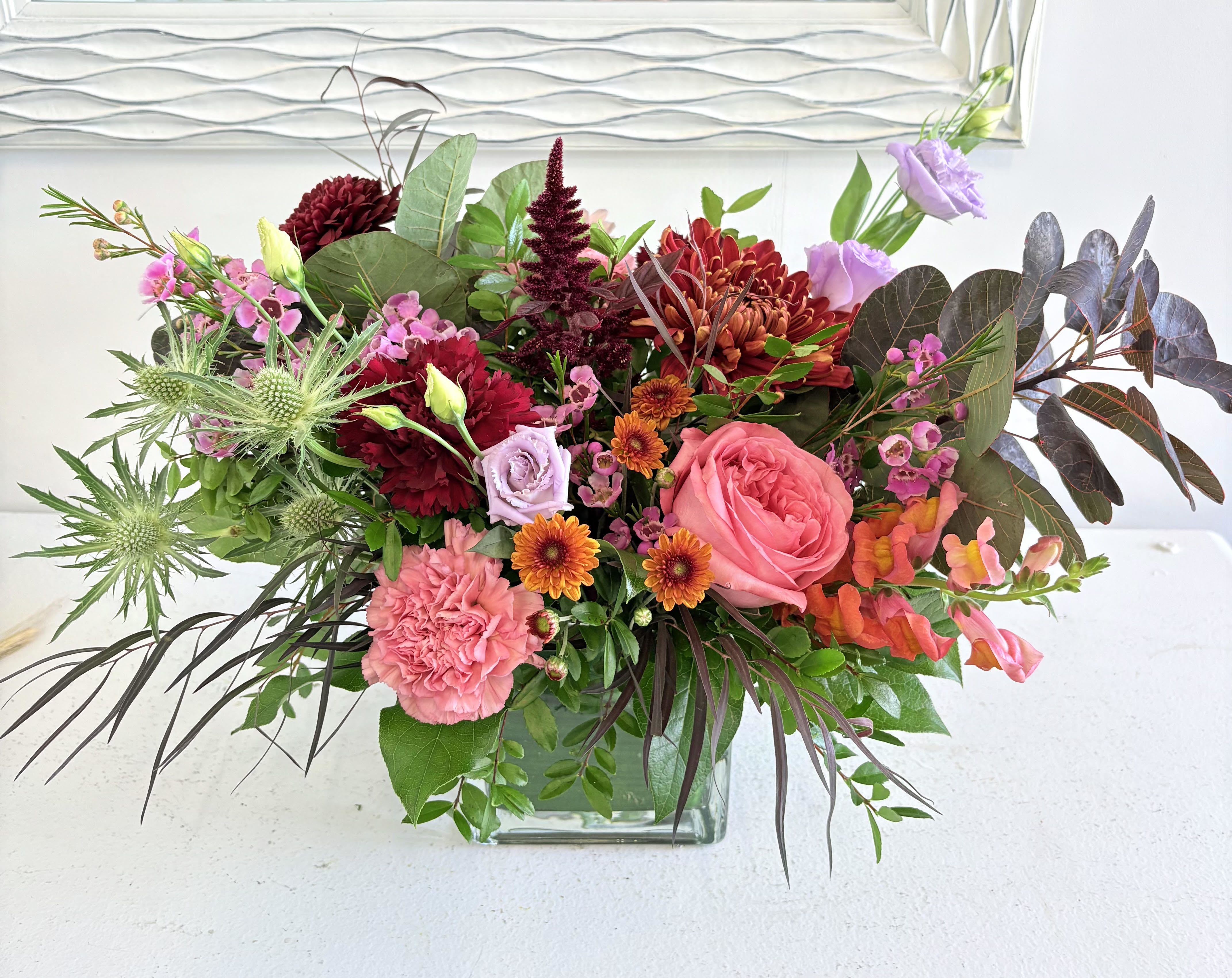 Crimson Glow - Elegant and full of depth, Crimson Glow captures the dramatic beauty of autumn in bloom. Lush roses, carnations, and mums in shades of burgundy, coral, and blush mingle with lavender accents, thistle, and textured foliage for a look that’s both bold and sophisticated. Arranged in a modern glass vase, this radiant design brings warmth, color, and artistry to any space — a striking statement for the season.